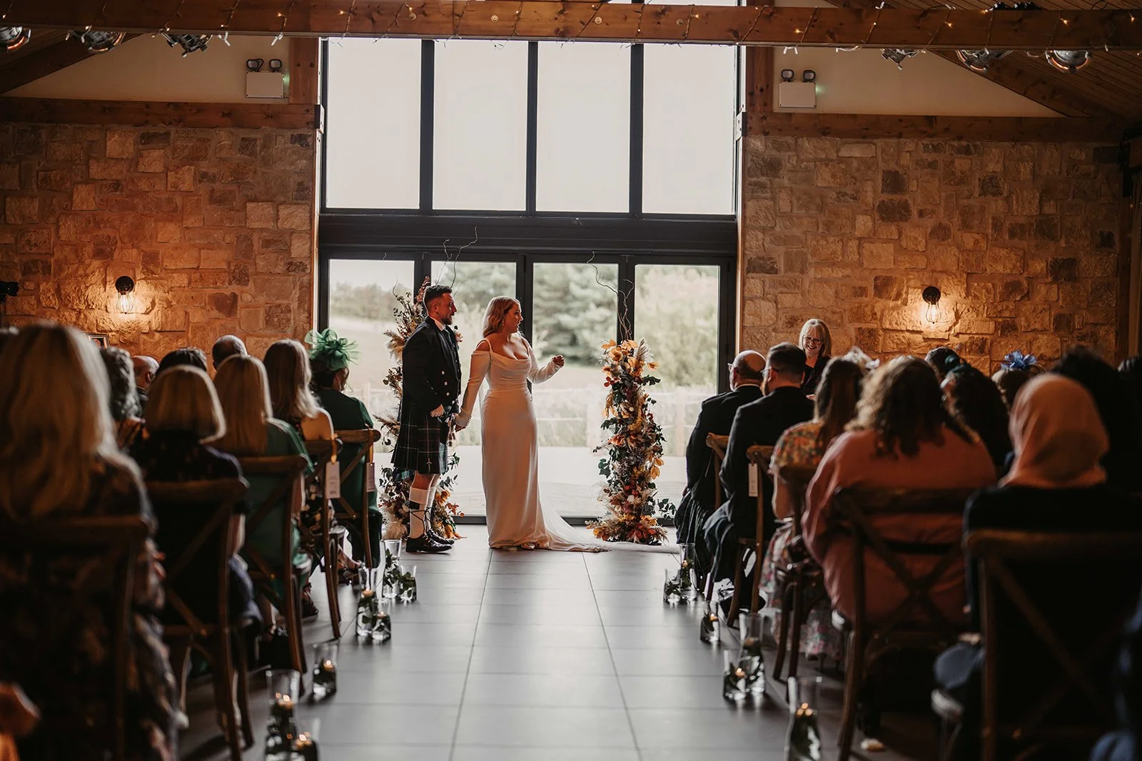 A wedding ceremony with a bride and groom standing holding hands in front of a large window. The bride is wearing a white dress, and the groom is in a kilt and jacket. Guests are seated on either side, with floral arrangements, and a woman officiating the ceremony is smiling.