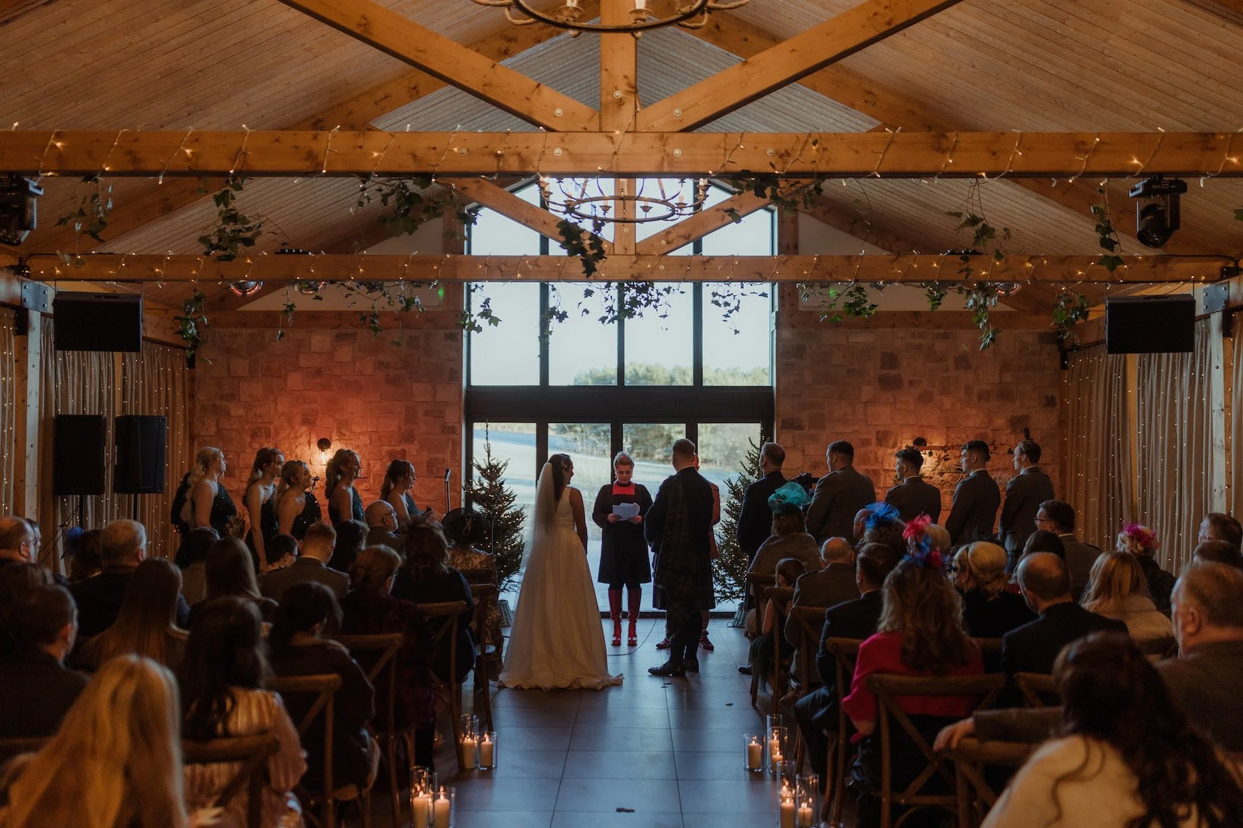 A wedding ceremony taking place inside a rustic wooden venue with high ceilings and large windows. The bride and groom stand at the altar in front of officiant, with bridesmaids to the left and groomsmen to the right. Guests are seated facing the couple, and candles are on the floor along the aisle. The room is decorated with fairy lights and greenery.