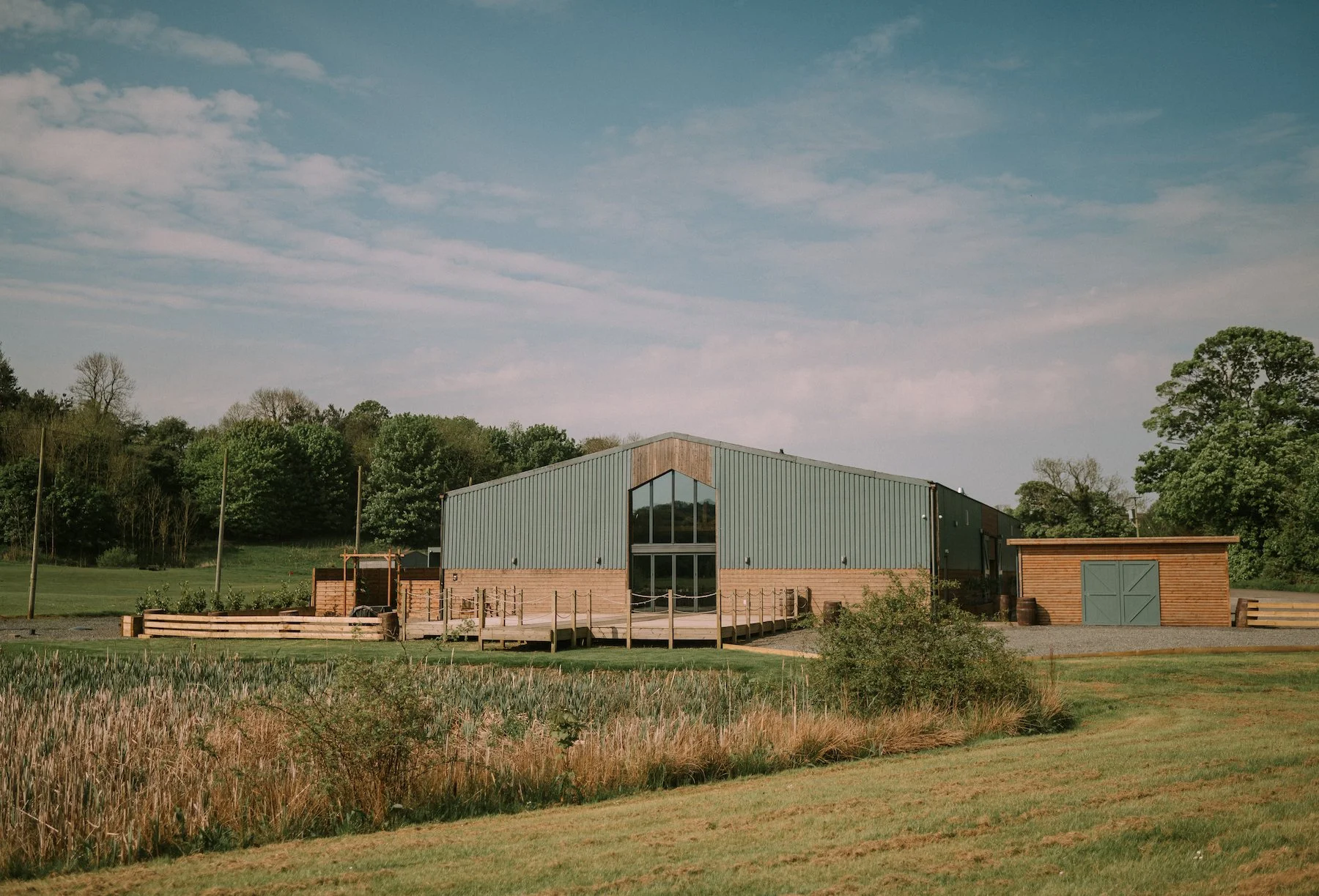 A modern building with large glass windows, surrounded by a landscaped area with grass, trees, and a wooden fence, under a partly cloudy sky.