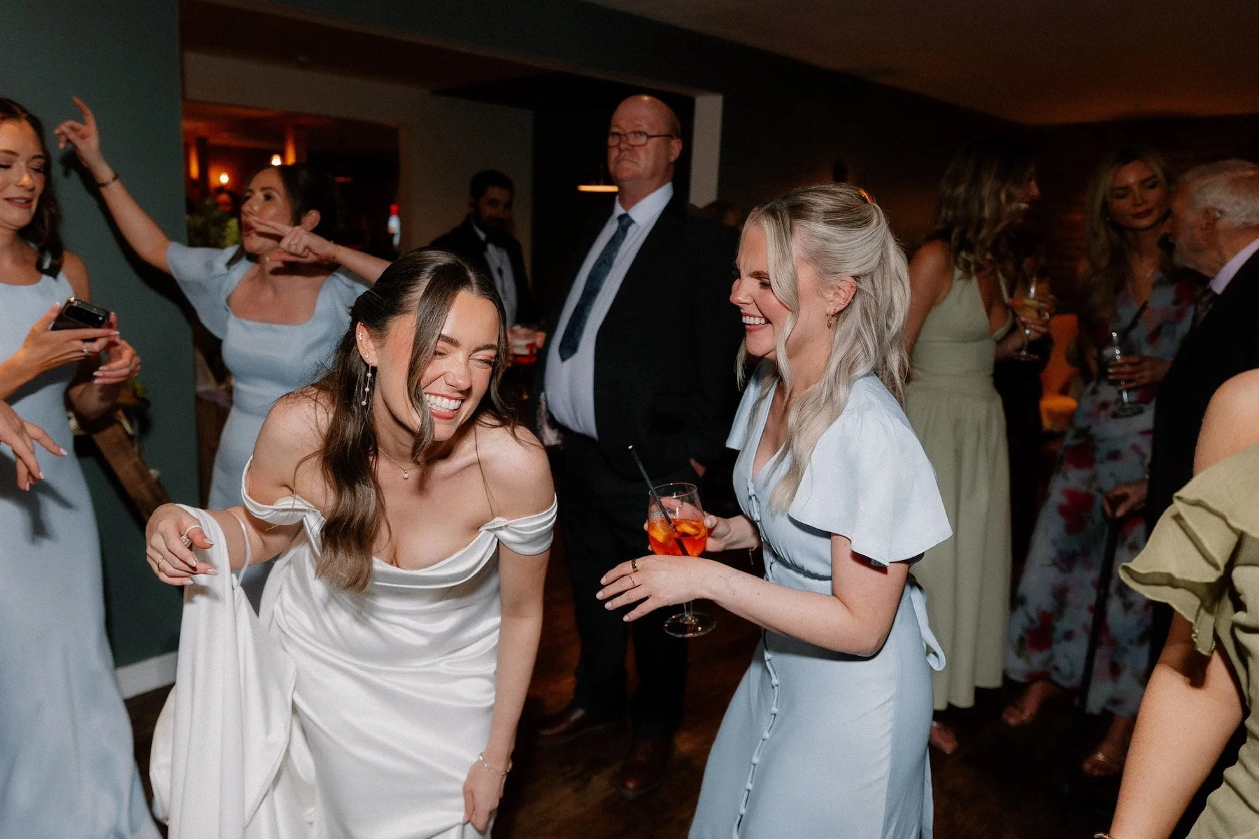 A group of people at a party, with two women in the foreground laughing and smiling while holding drinks, surrounded by other guests.