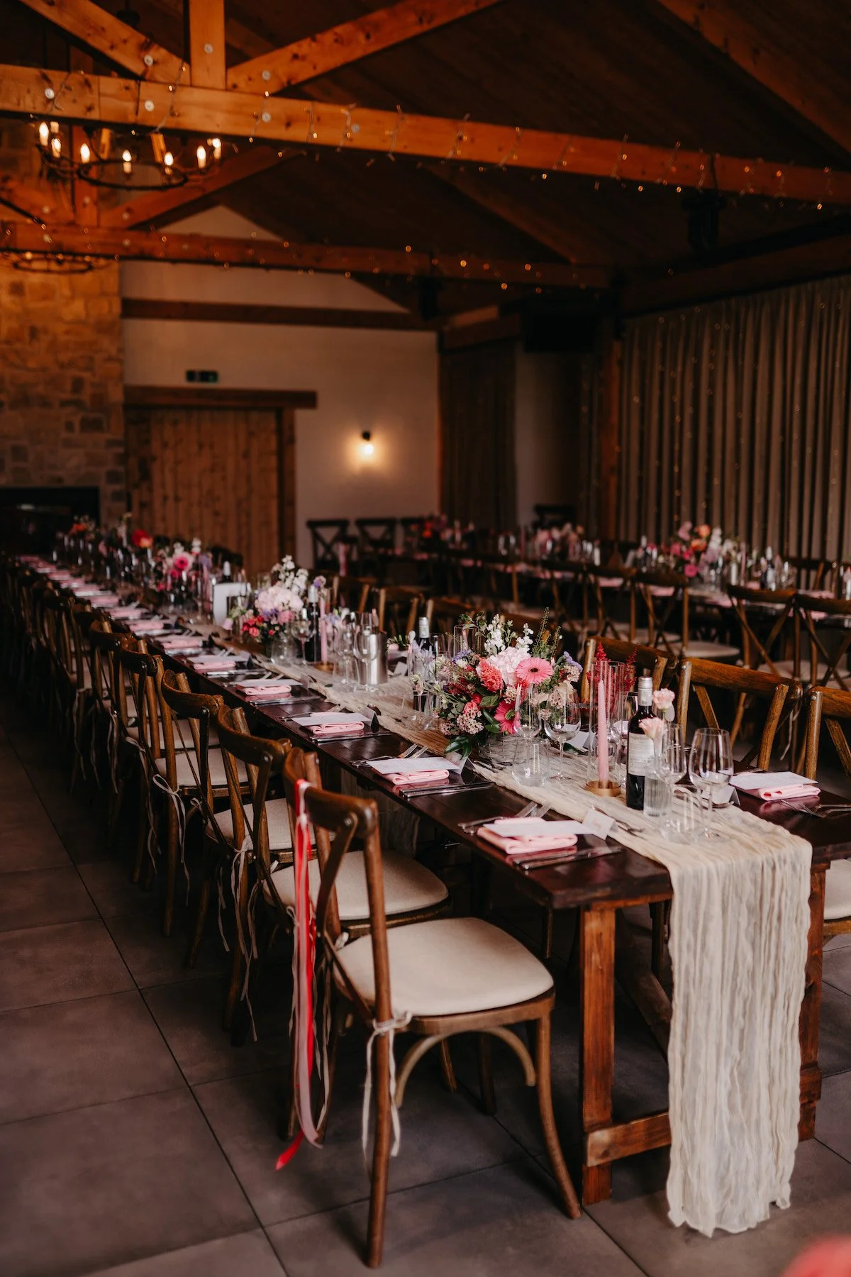 An elegant wedding reception table decorated with pink and white flowers, candles, glasses, and pink napkins in a rustic wooden hall with string lights.