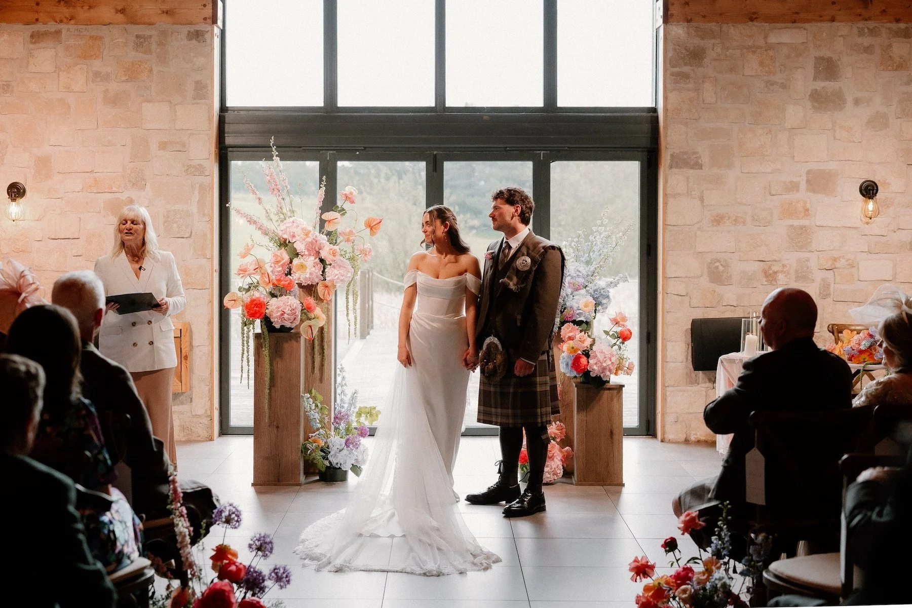 A wedding ceremony with a bride and groom holding hands in front of a minister, surrounded by floral arrangements, with guests sitting and watching inside a modern venue with large windows and stone walls.