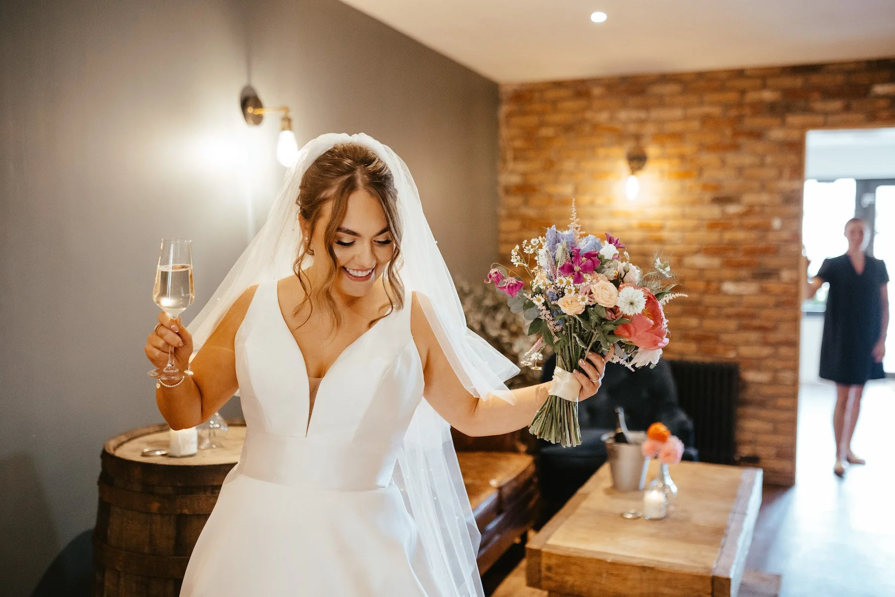 A bride in a white wedding dress holding a bouquet of colorful flowers and a glass of champagne, smiling indoors with exposed brick wall and a woman in black dress in the background.