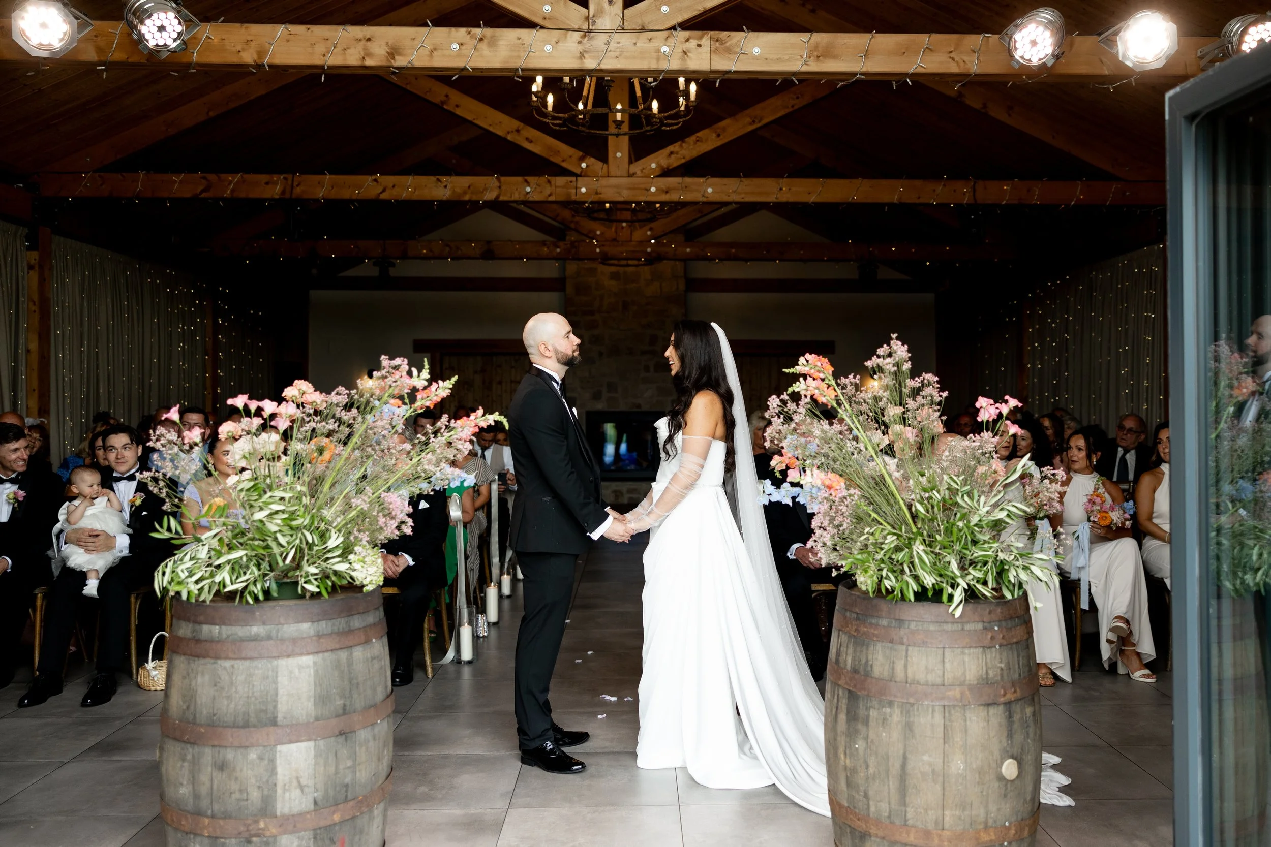 A bride and groom stand facing each other during their wedding ceremony inside a decorated venue, surrounded by guests, with floral arrangements and warm lighting in a rustic setting.
