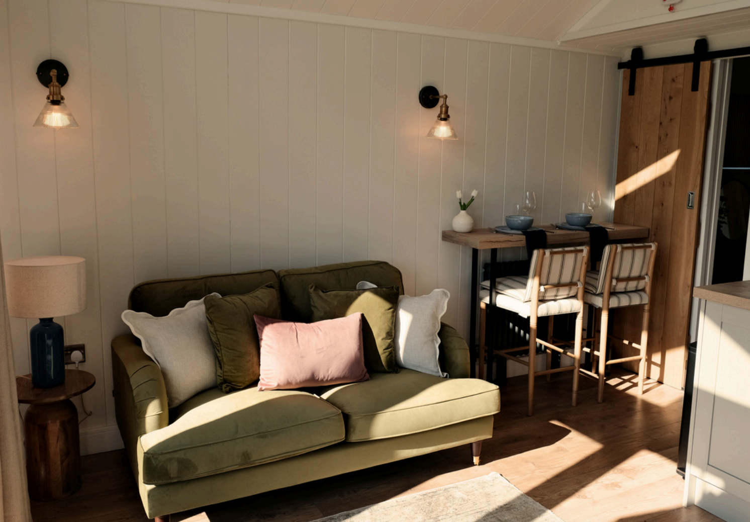 Living room with a green sofa, pillows, a small wooden side table with a blue lamp, wall-mounted lights, a small dining table with two chairs, wine glasses, bowls, and a vase with flowers, in front of a white wood-paneled wall and a wooden sliding door.