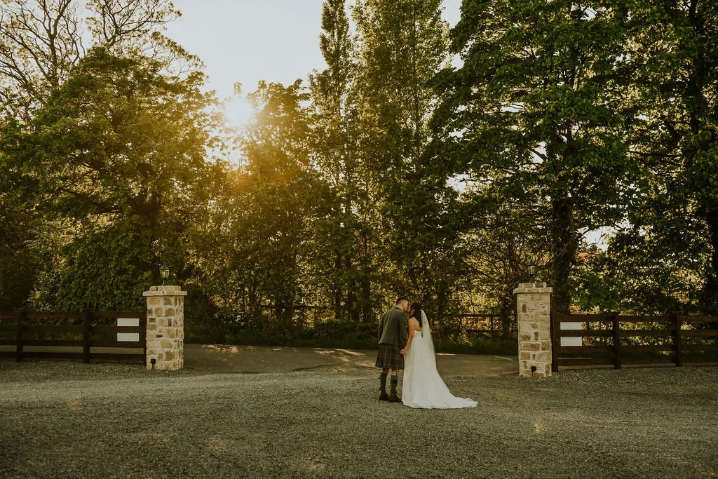 A bride and groom stand close together on a gravel pathway, surrounded by trees, with sunlight filtering through the foliage.