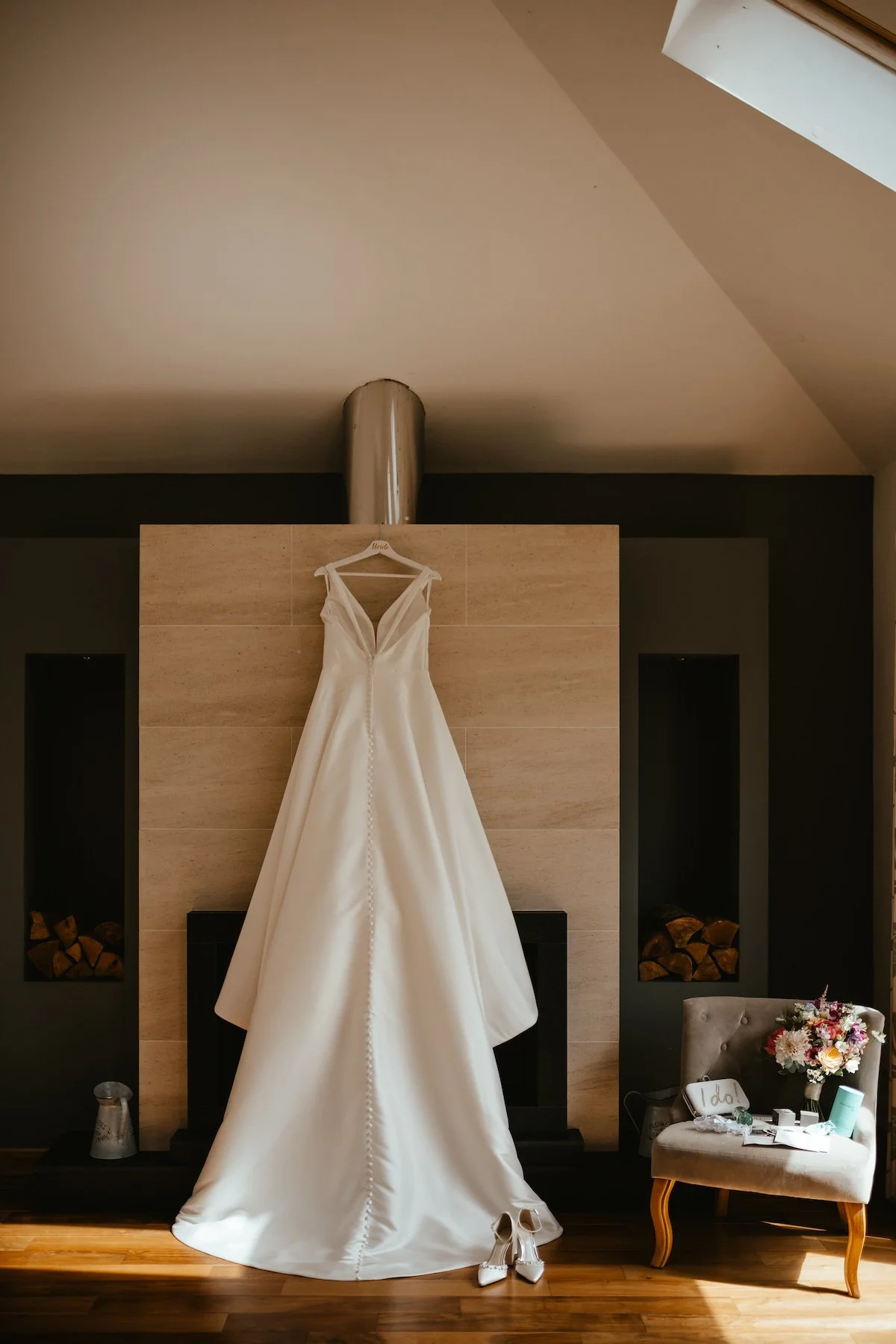 A wedding dress hanging on a hanger above a fireplace, with white high heels placed underneath on a wooden floor. To the right, a cozy armchair holds a bouquet of flowers and various small items.