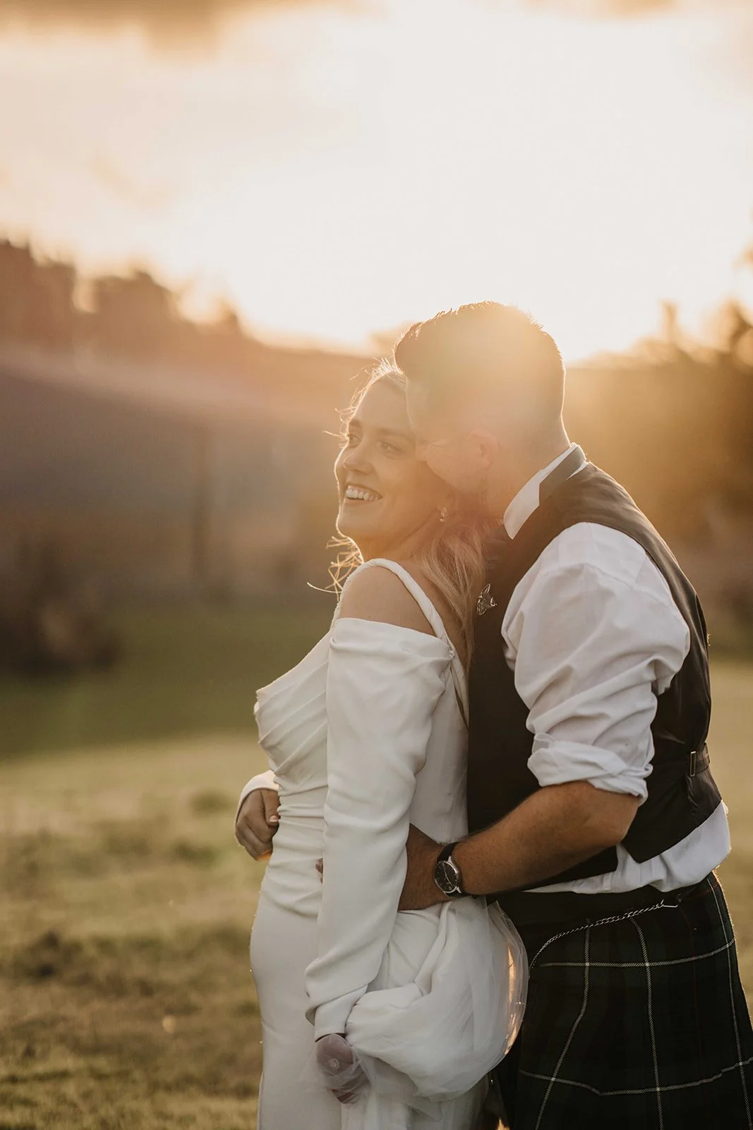 A couple hugging outdoors at sunset, smiling, the woman wearing a white dress and the man in a shirt with rolled-up sleeves and a vest.