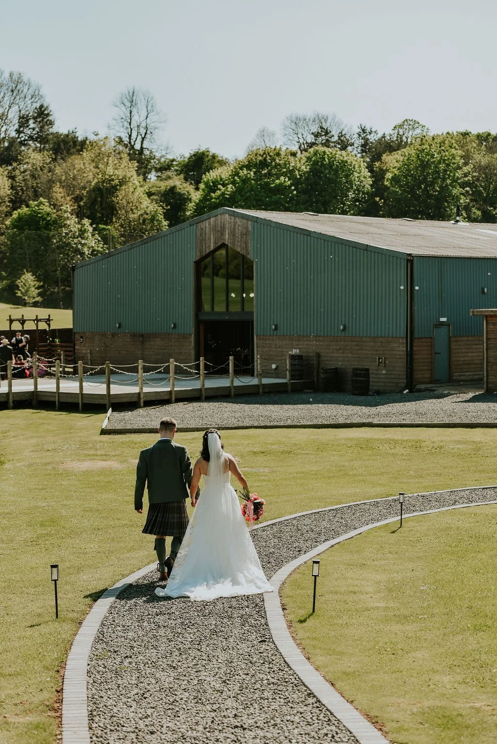 Bride and groom walking hand-in-hand on a curved stone pathway towards a rustic green barn, with a backdrop of trees, on a sunny day.