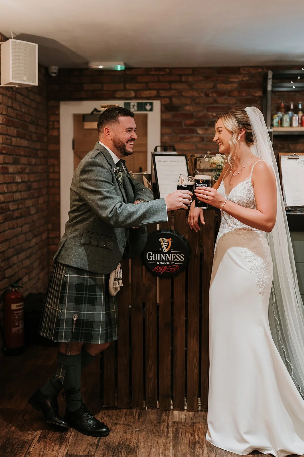 A bride and groom smiling and clinking Guinness drinks at their wedding reception in a cozy, rustic venue with brick walls.