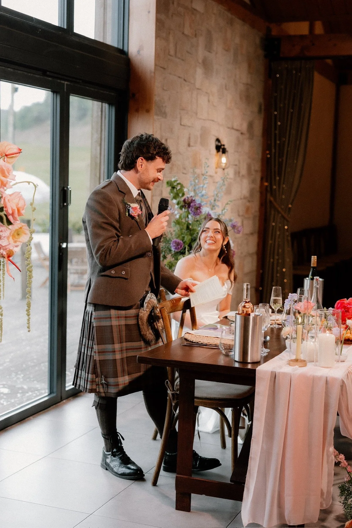A man in traditional Scottish attire giving a speech at a wedding reception, with a woman in a white dress smiling and seated at a decorated table with floral arrangements and candles.