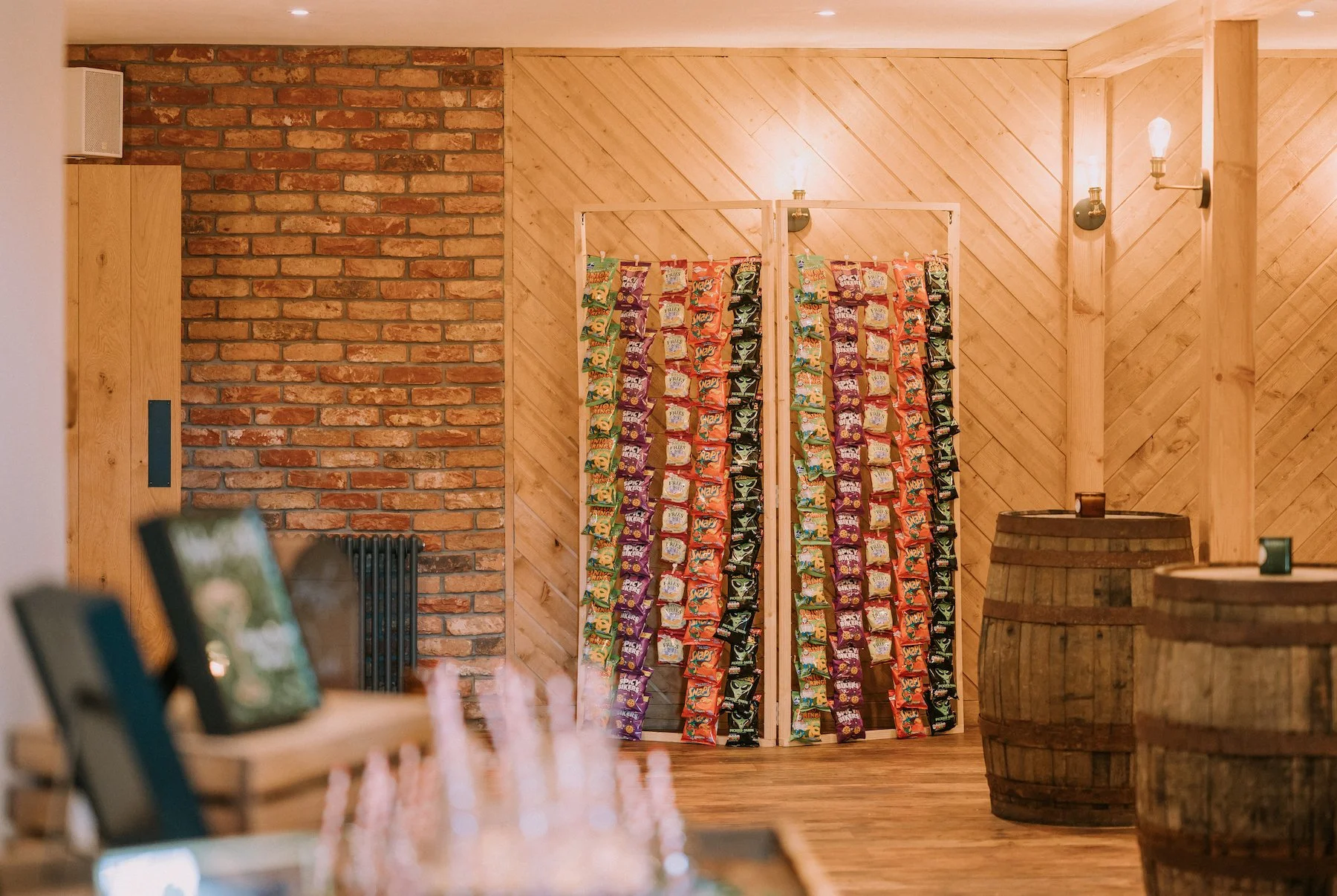 Display of various colorful snack bags hanging on a wooden frame, set against a rustic brick and wood-paneled wall, with barrels nearby in a cozy indoor setting.