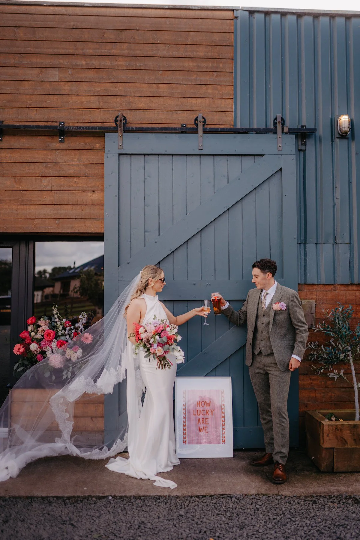 A bride and groom celebrating with drinks outside a rustic building with wooden and metal siding. The bride is holding a bouquet of pink and white flowers and wearing a white gown with a long veil, while the groom is dressed in a gray suit with a flower boutonniere. There is a sign at their feet that reads "How Lucky Are We" and a floral arrangement nearby.