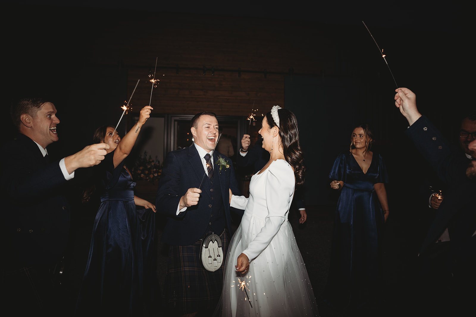 A group of people celebrating a wedding with sparklers in a dark room, with the bride and groom smiling at each other in the center.