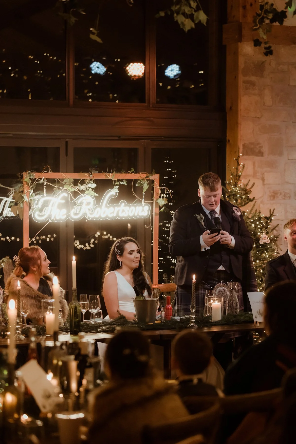 A man in traditional Scottish attire giving a speech at a wedding reception, seated at a decorated table with candles, surrounded by guests. The backdrop has a sign that says 'The Robertsons' and festive Christmas decorations.