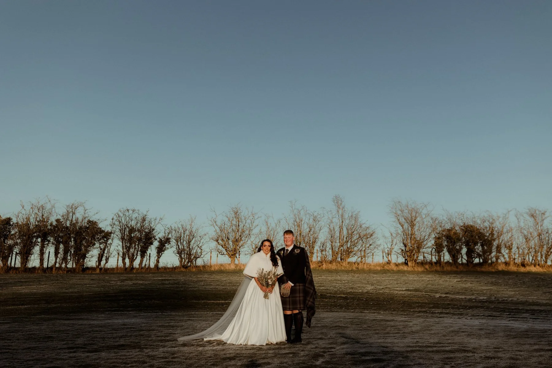 A bride and groom standing outdoors in a wide open field with leafless trees in the background under a clear sky at sunset.
