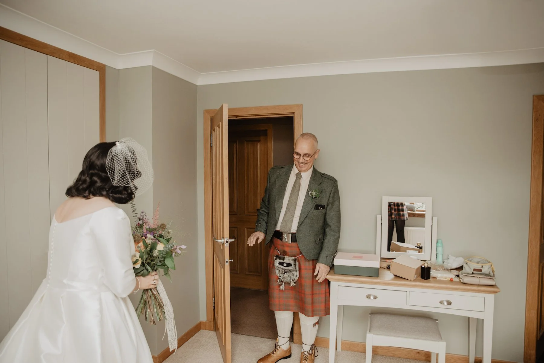 A bride holding a bouquet standing in a room, facing a man dressed in a kilt and blazer, smiling.