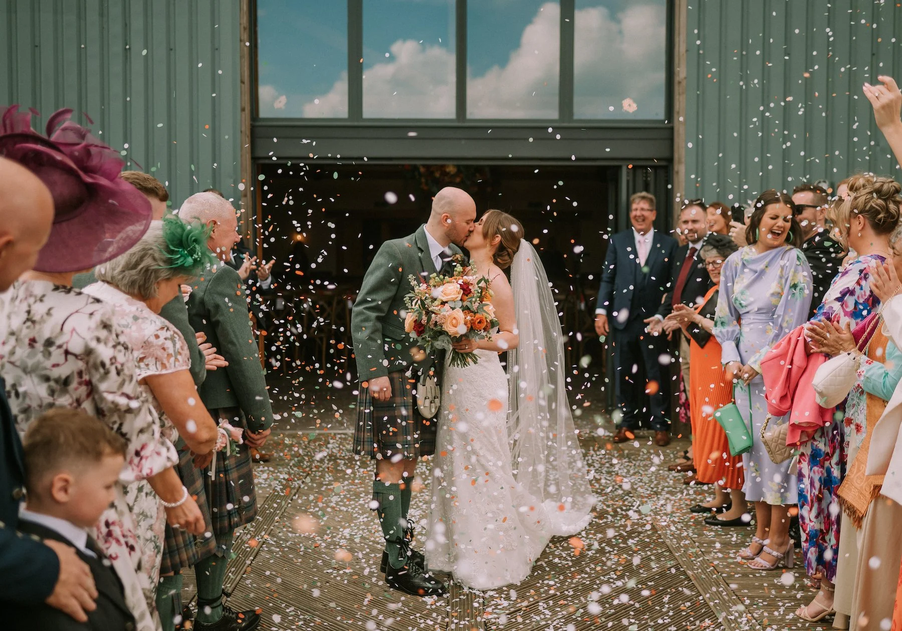 Bald man in traditional Scottish kilt and jacket kisses a bride holding a bouquet of flowers, as guests throw confetti outside a building with large windows behind them.