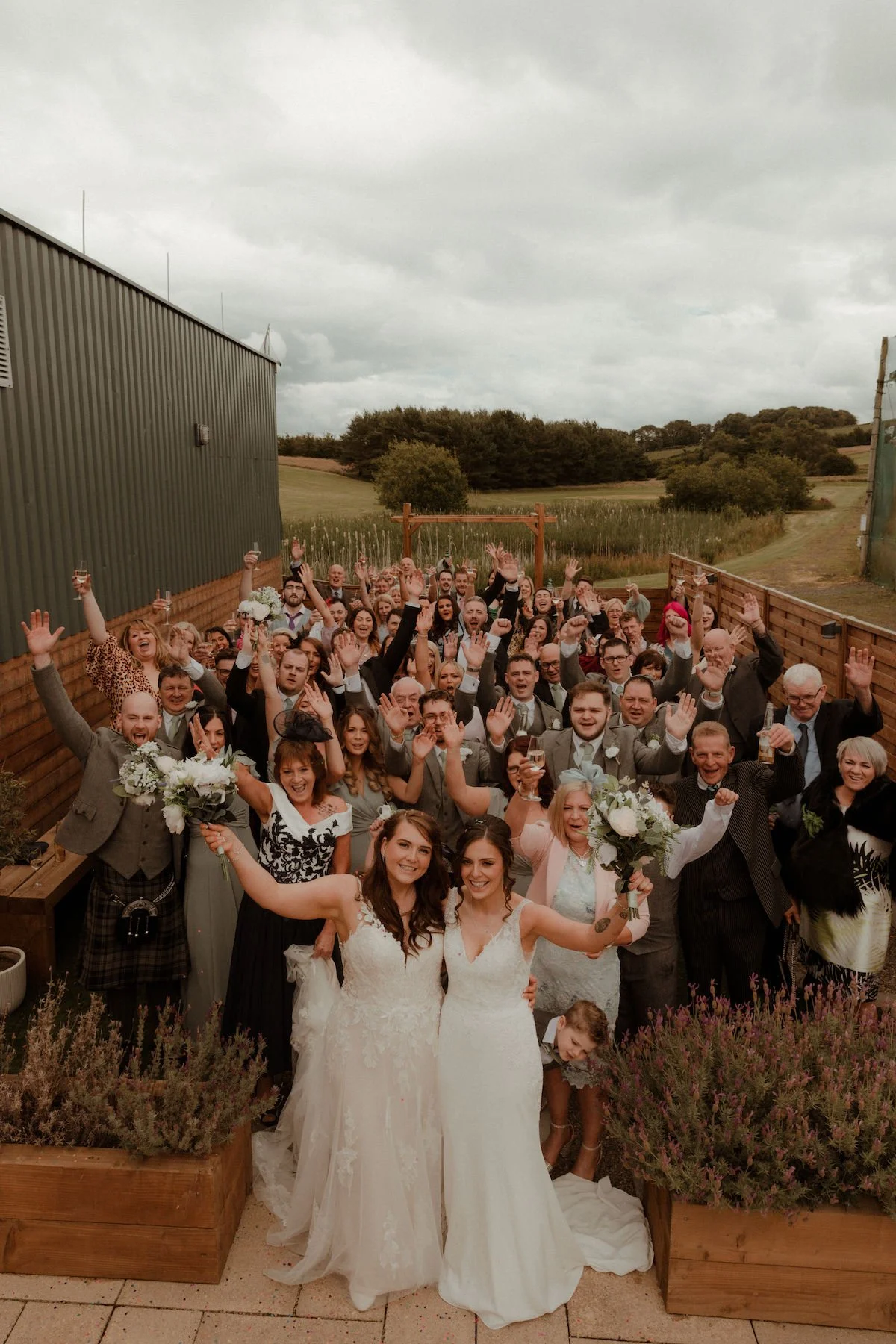 A large group of wedding guests celebrating outdoors, with two brides in wedding dresses at the front, holding bouquets, and many guests raising their hands and glasses in celebration.