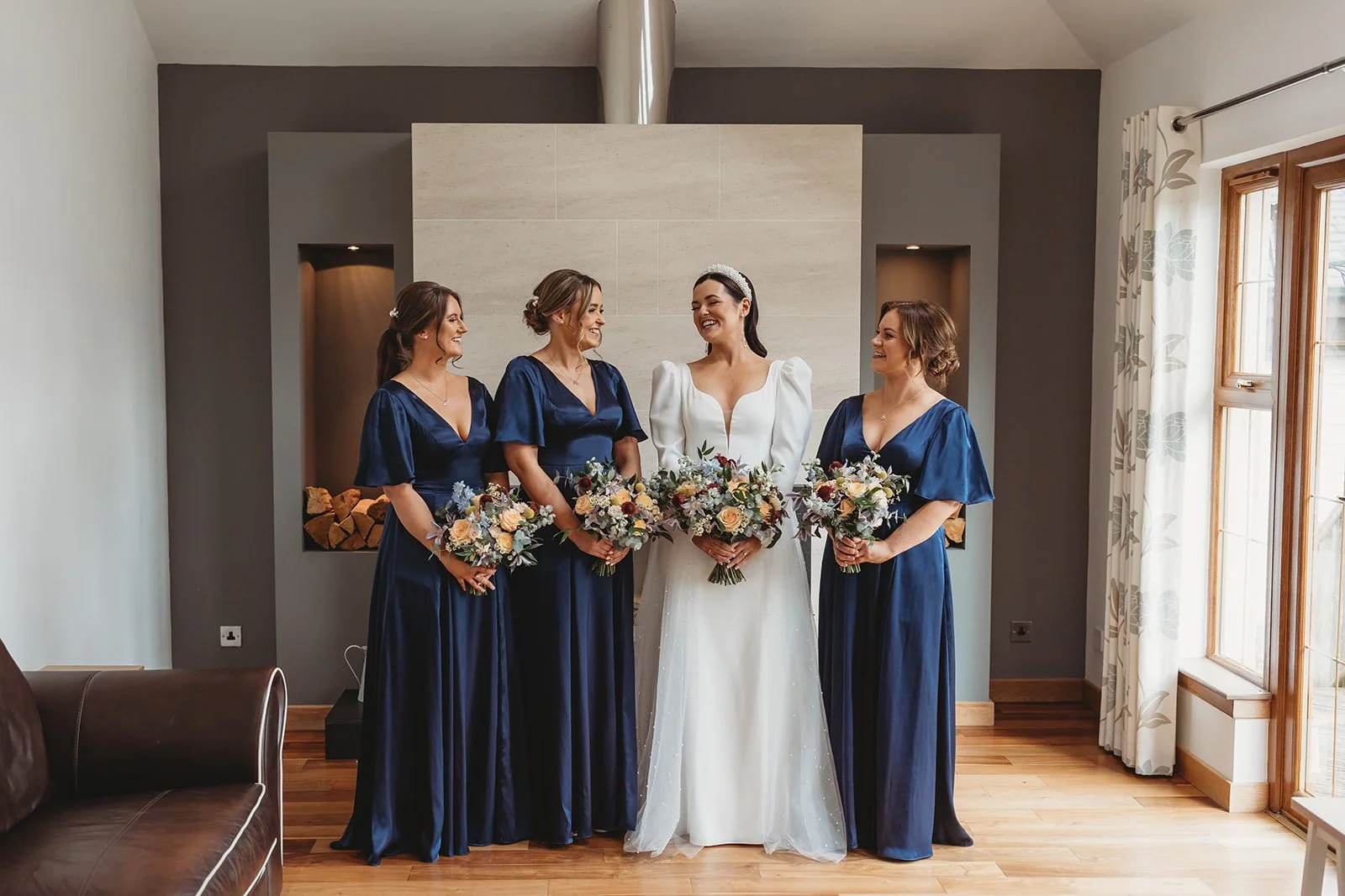 A bride and three bridesmaids stand together indoors, smiling and holding bouquets. The bride wears a white gown with puffed sleeves and a tiara, while the bridesmaids wear matching navy blue dresses.