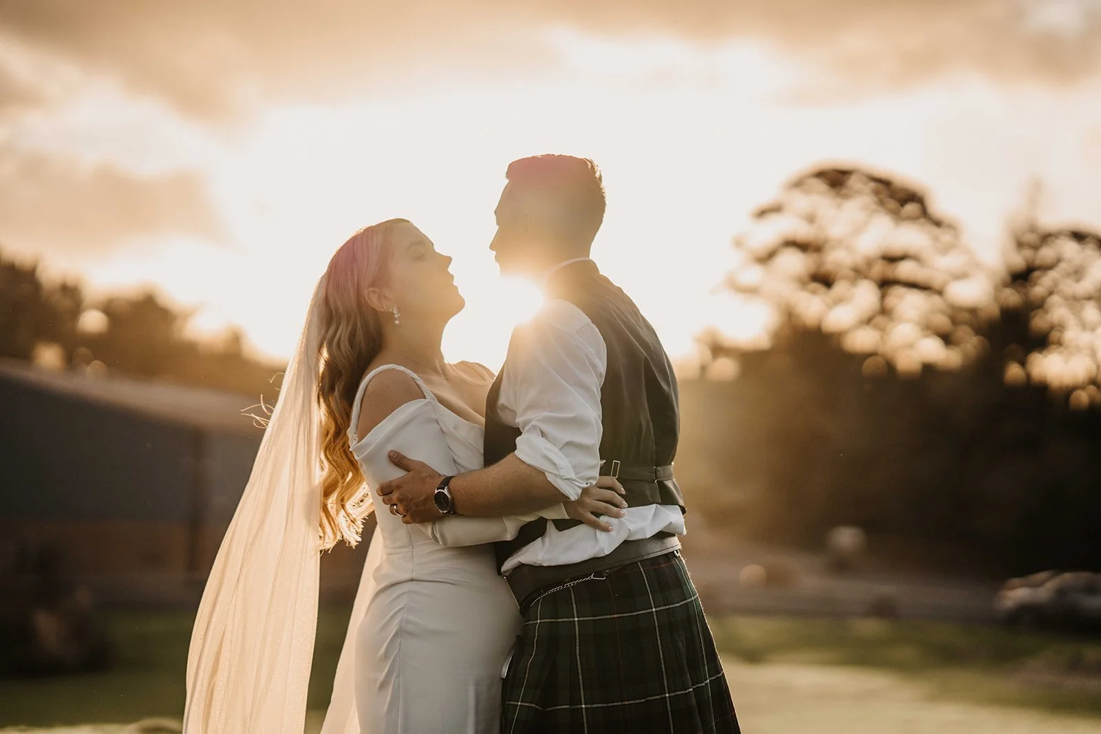 A couple dressed in wedding attire standing close and embracing outdoors at sunset, with the sun shining behind them and trees in the background.