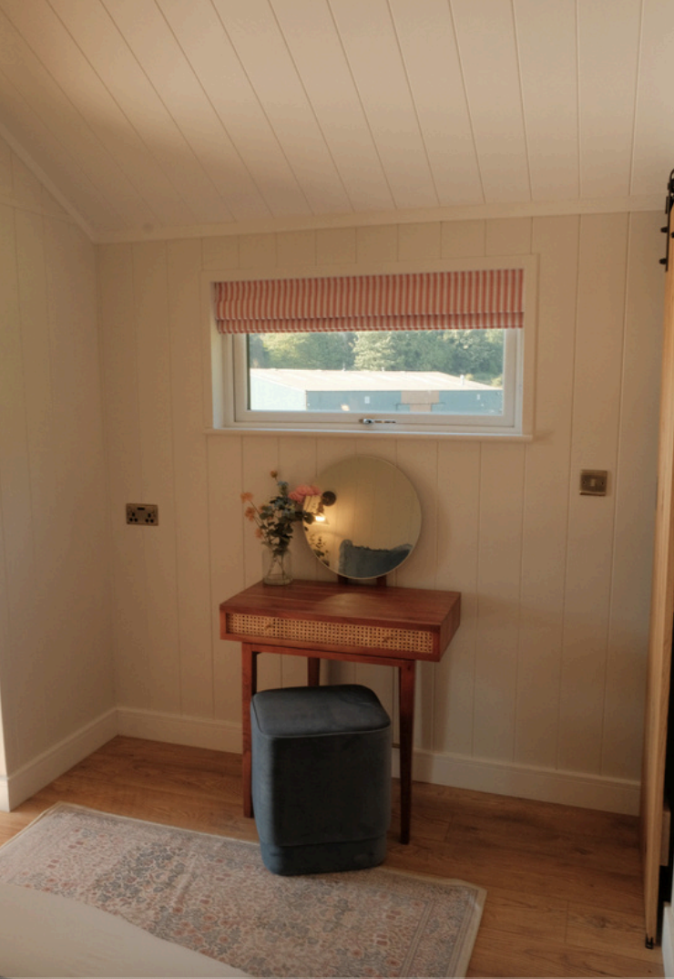 A cozy corner with a small wooden vanity table, a round mirror, a vase with flowers, a dark upholstered stool, a window with a red-striped curtain, and a decorative rug on wooden flooring.