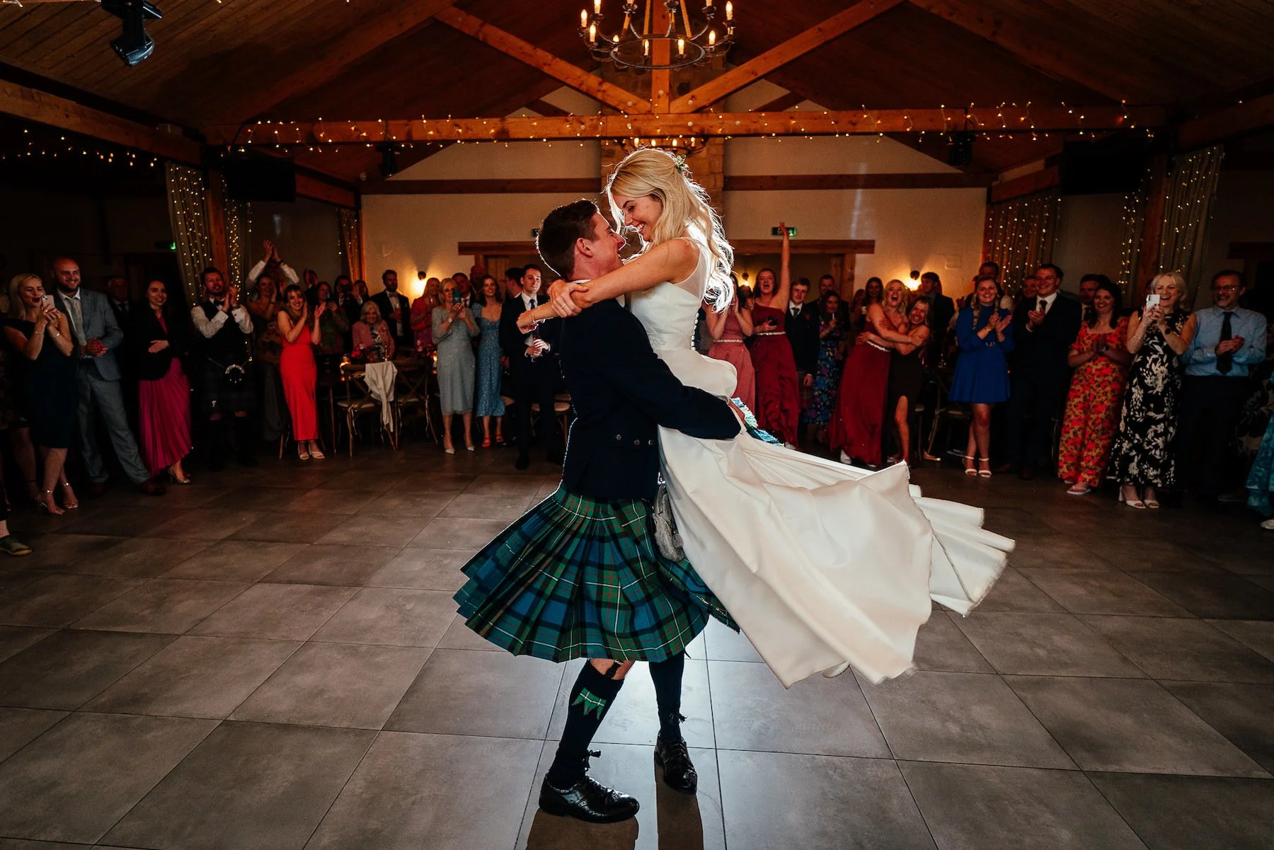 A bride and groom dancing at their wedding reception, with the groom in a kilt lifting the bride in a white wedding gown, while guests watch and cheer in a warmly lit rustic venue decorated with string lights and chandeliers.