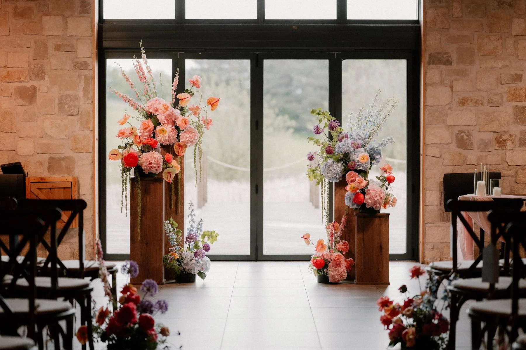 Indoor venue decorated with tall floral arrangements featuring pink, purple, and red flowers on wooden stands, set in front of glass doors.
