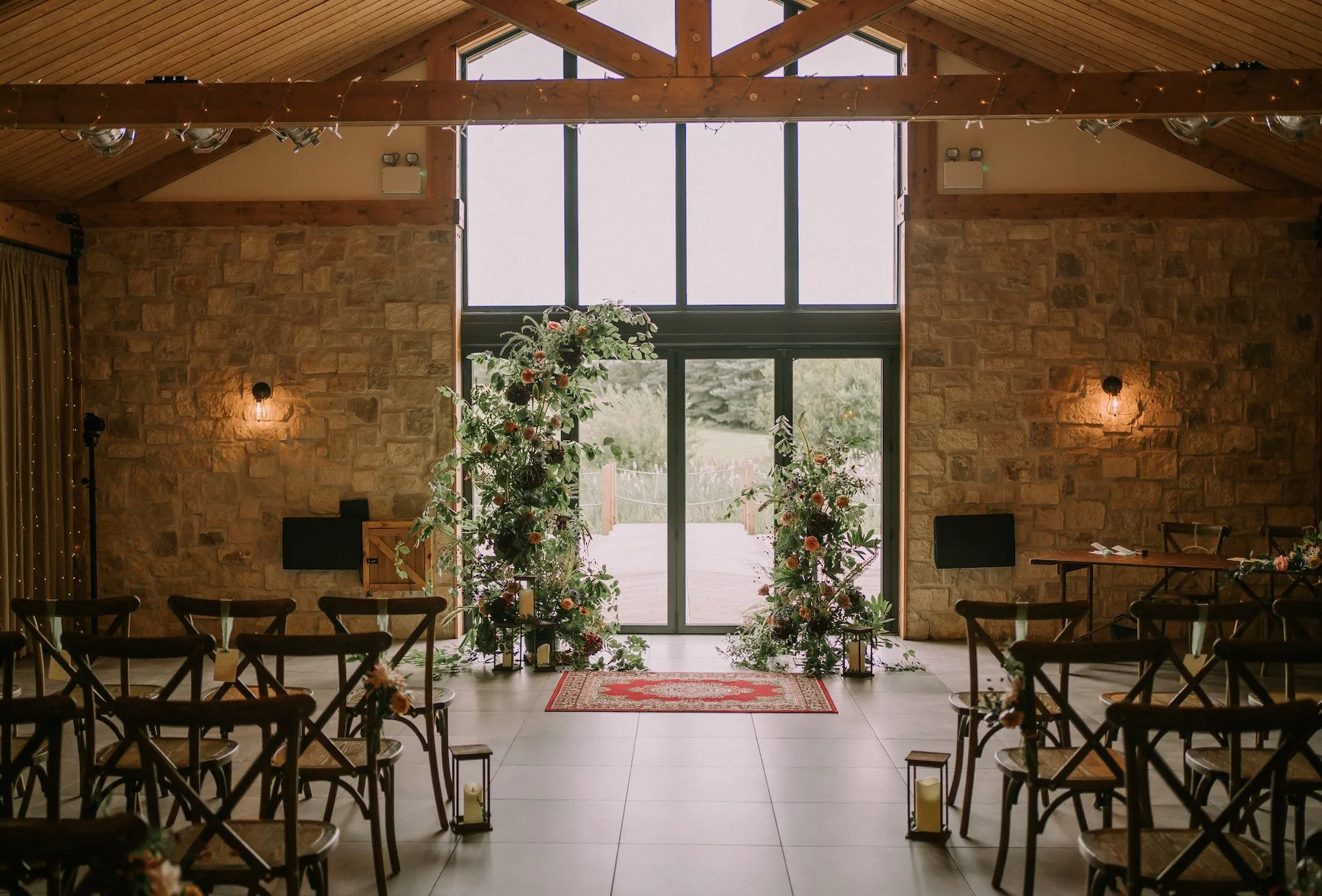 Wedding ceremony setup inside a rustic stone and wood venue with chairs arranged on either side of an aisle decorated with flowers and lanterns, leading to a large window and glass door at the front with outdoor scenery visible beyond.