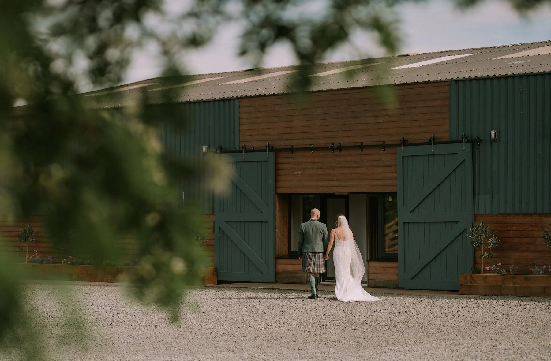 A bride and groom walking into a rustic barn-style venue, framed by blurred green foliage in the foreground.