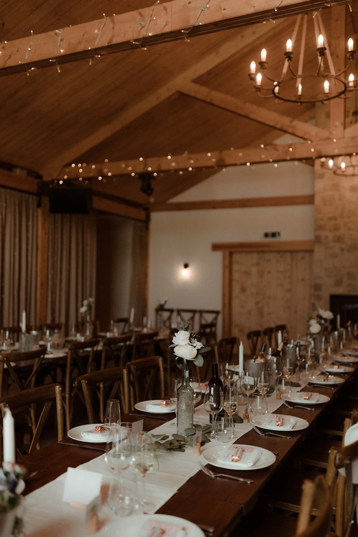 Interior of a rustic wedding reception with a long table decorated with white flowers, candles, and wine bottles, featuring wooden chairs, exposed brick wall, wooden ceiling beams, and hanging string lights and chandeliers.