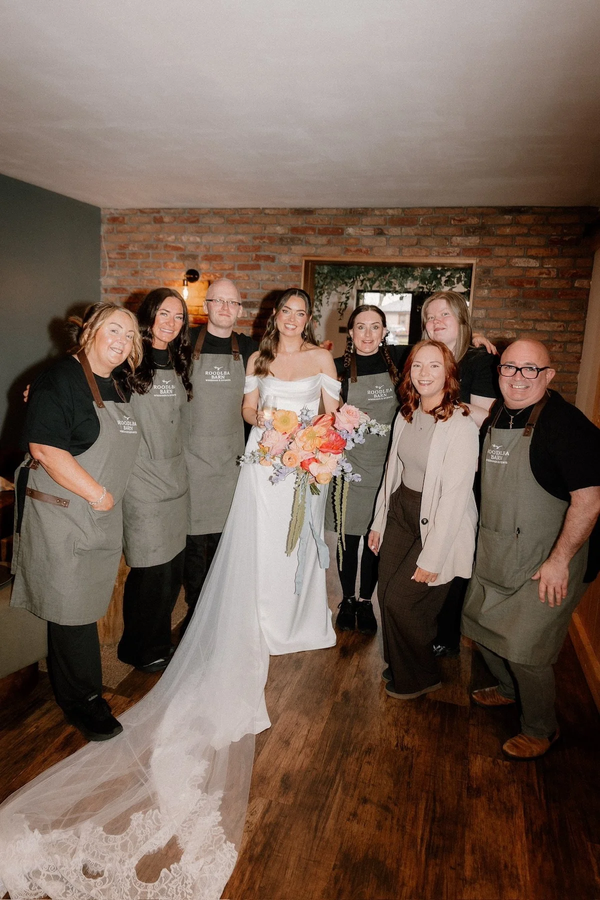 A group of nine people, including a bride in a white wedding dress holding a bouquet, posing for a photo in a rustic indoor setting with a brick wall background. Five of them are wearing aprons.
