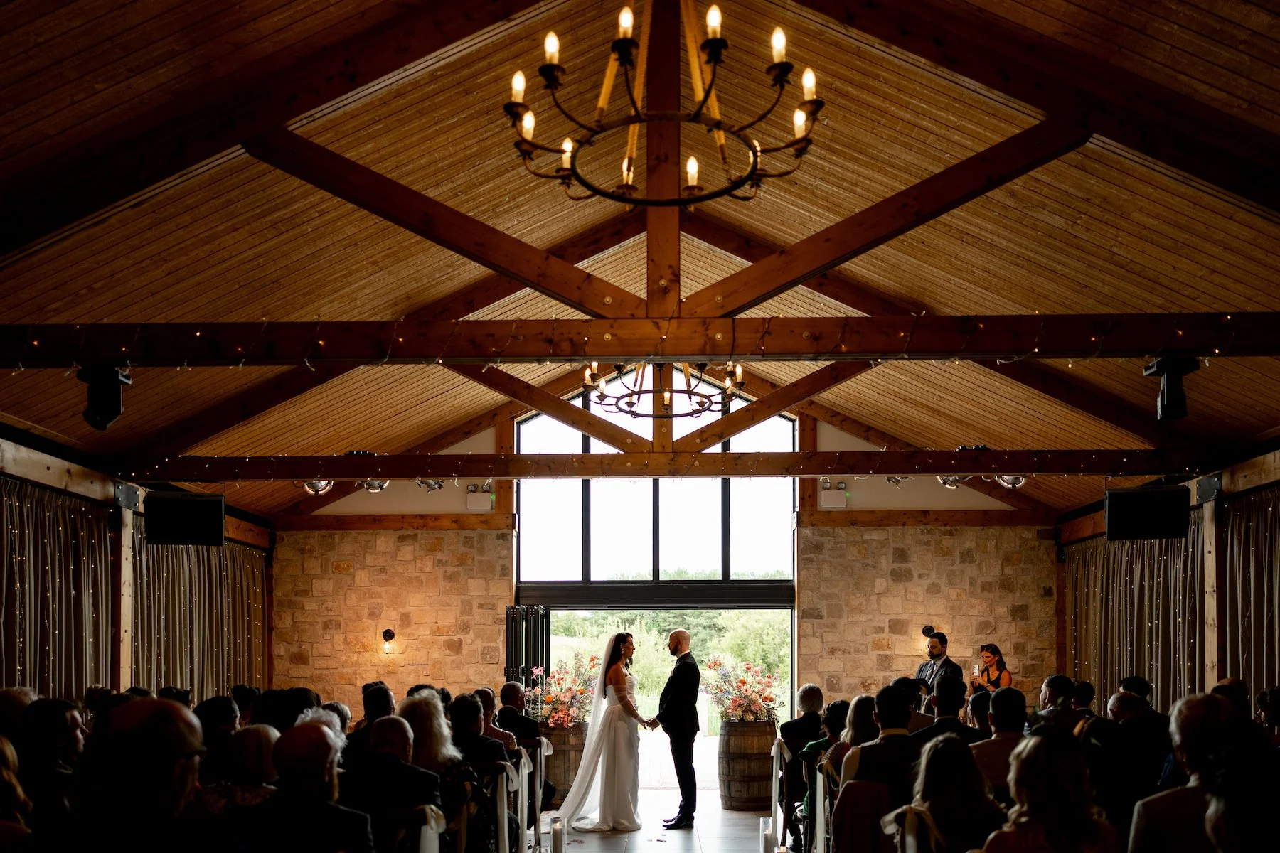 A bride and groom standing together, holding hands, in a rustic wedding ceremony inside a wooden venue with string lights and chandeliers, with guests seated around them.