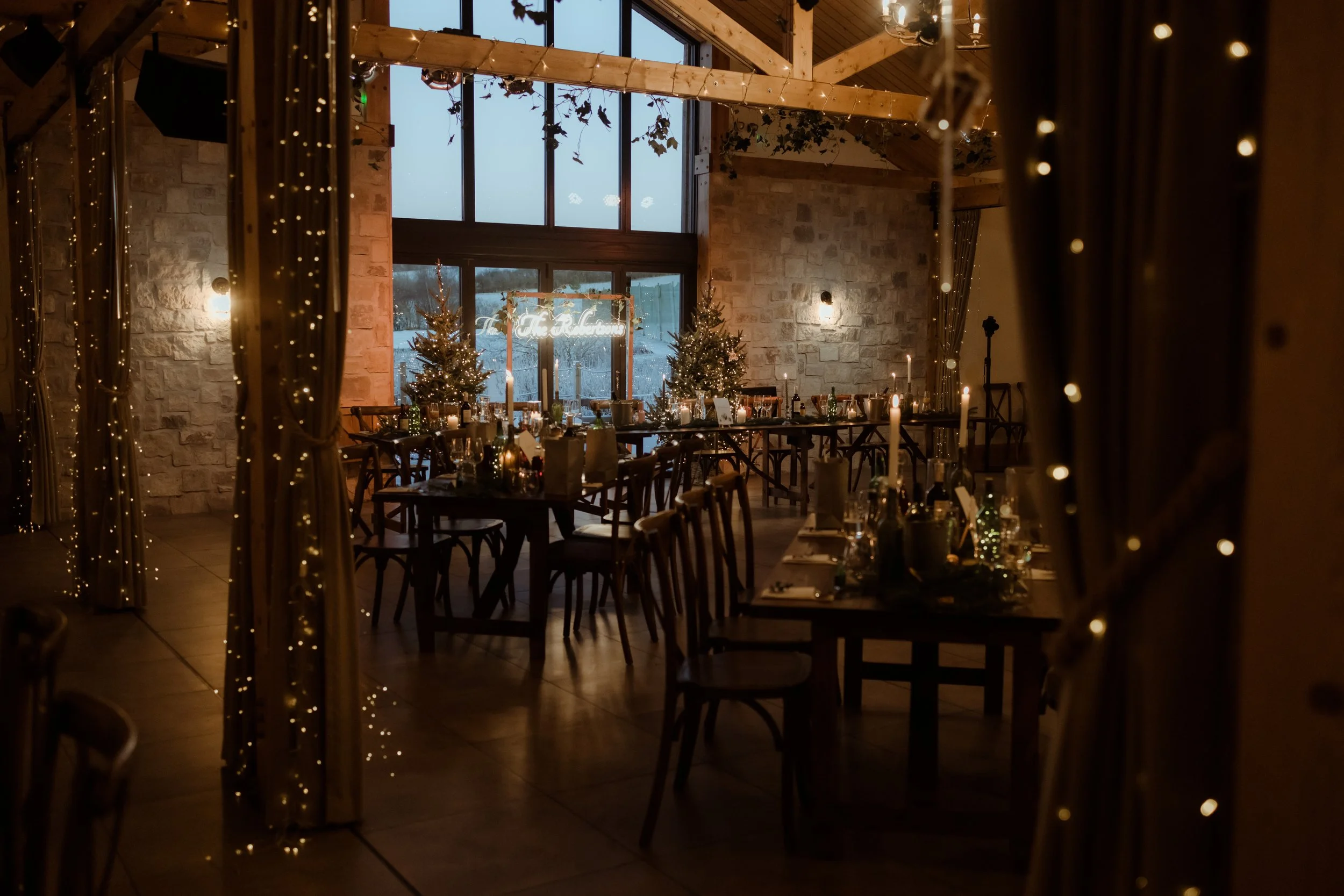 A warmly lit indoor celebration space decorated with Christmas trees, fairy lights, and candles, with tables set for guests and a large window showing an outdoor view.
