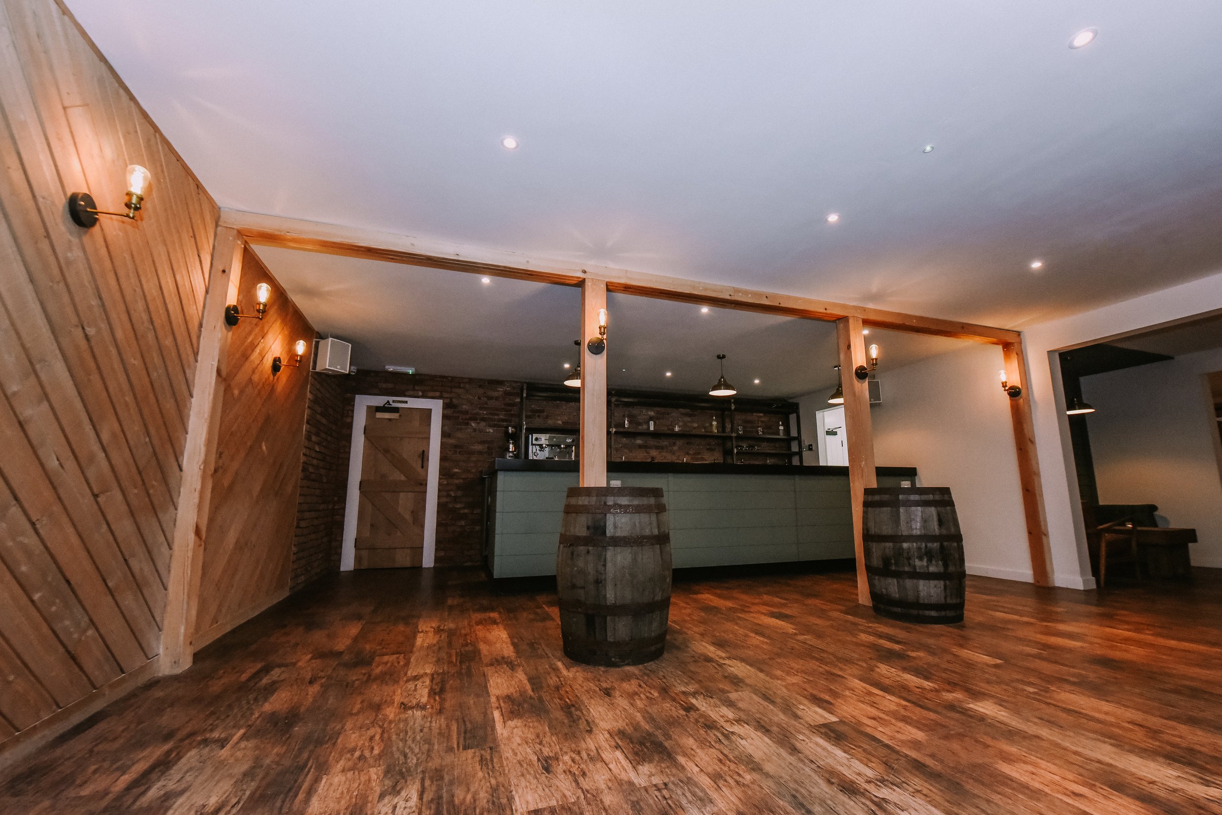 Interior of a rustic bar or restaurant with wooden floors, walls, and barrels, with a bar counter in the background and ceiling lights.