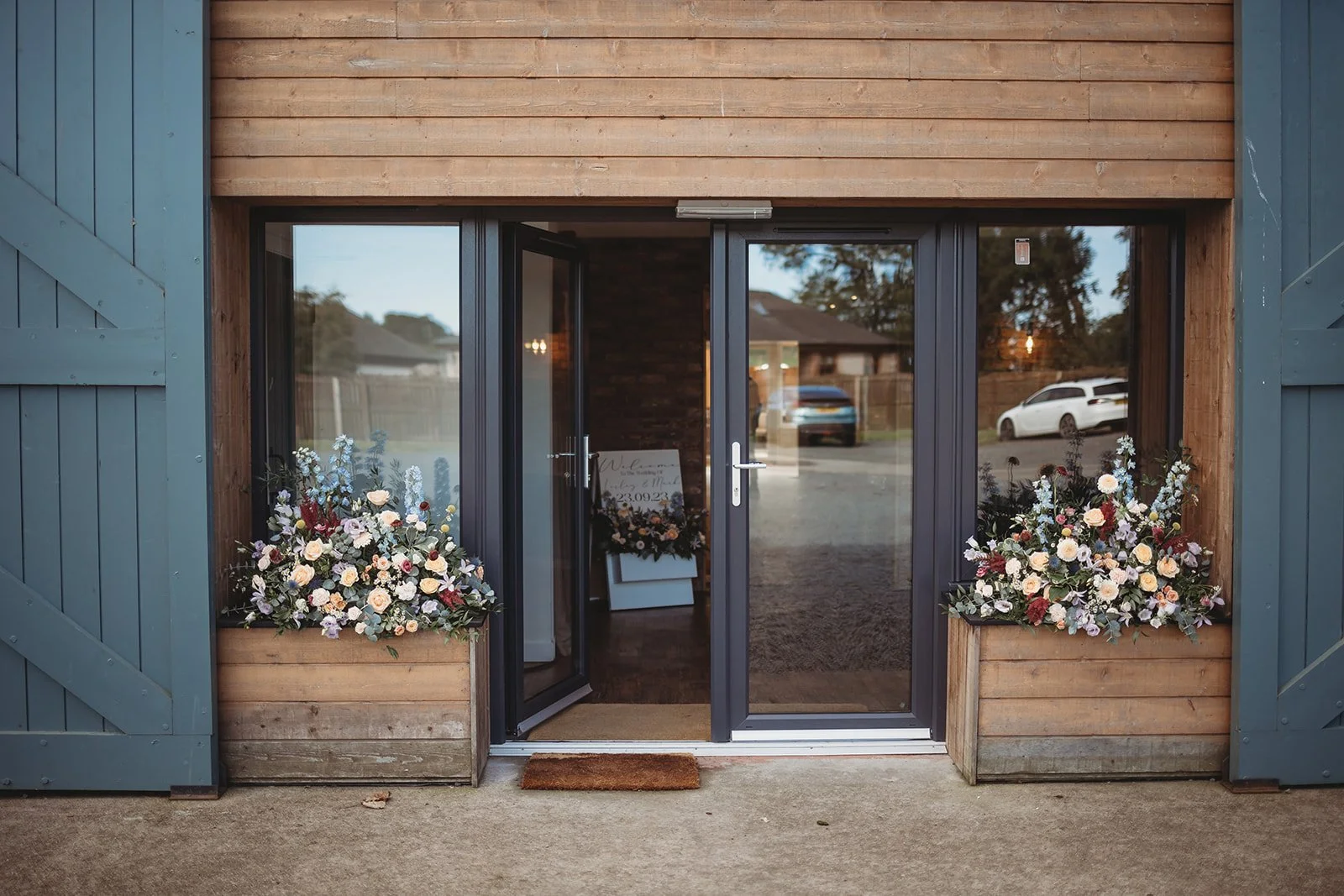 Entrance to a building decorated with two large flower arrangements on either side of the glass door, a welcome sign inside, and a brown doormat outside the door.