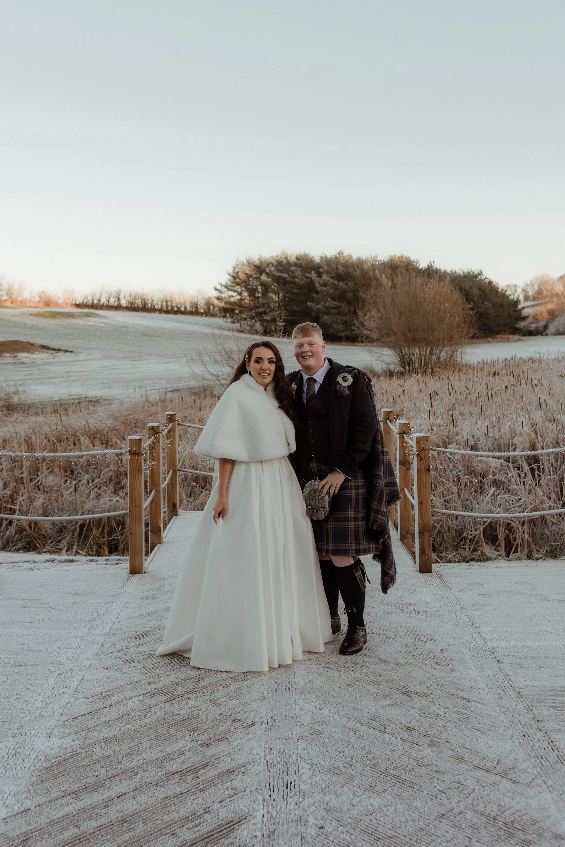 A couple dressed in wedding attire standing on a wooden bridge outdoors in a winter setting, with snow on the ground and trees in the background.