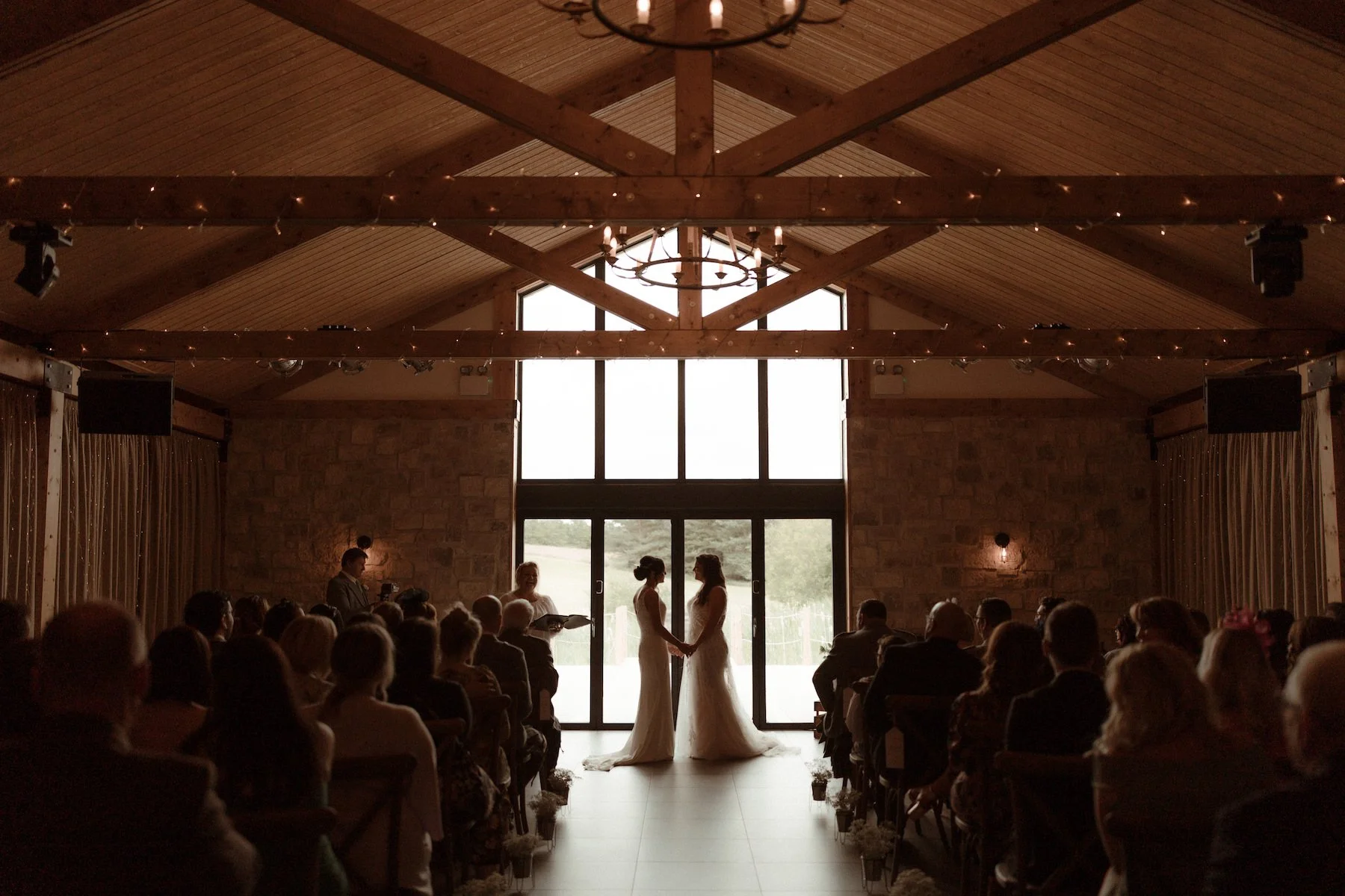 Two women in wedding dresses holding hands and facing each other in front of a large window during a wedding ceremony in a rustic venue with exposed wooden beams, chandeliers, and an audience watching.