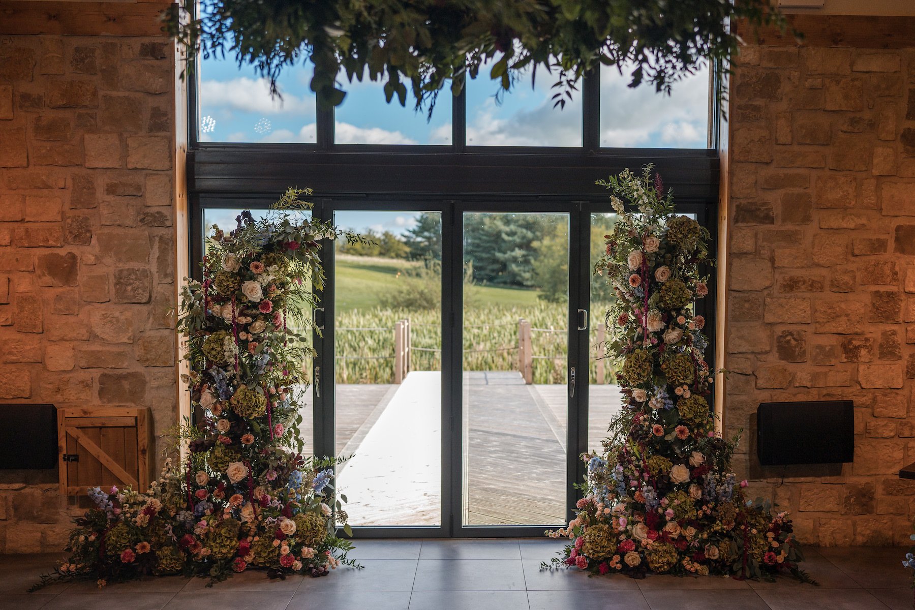 Decorative floral archway with various flowers and greenery stands in front of glass doors leading to an outdoor area with a wooden deck and green landscape, inside a building with brick walls and a wood ceiling.