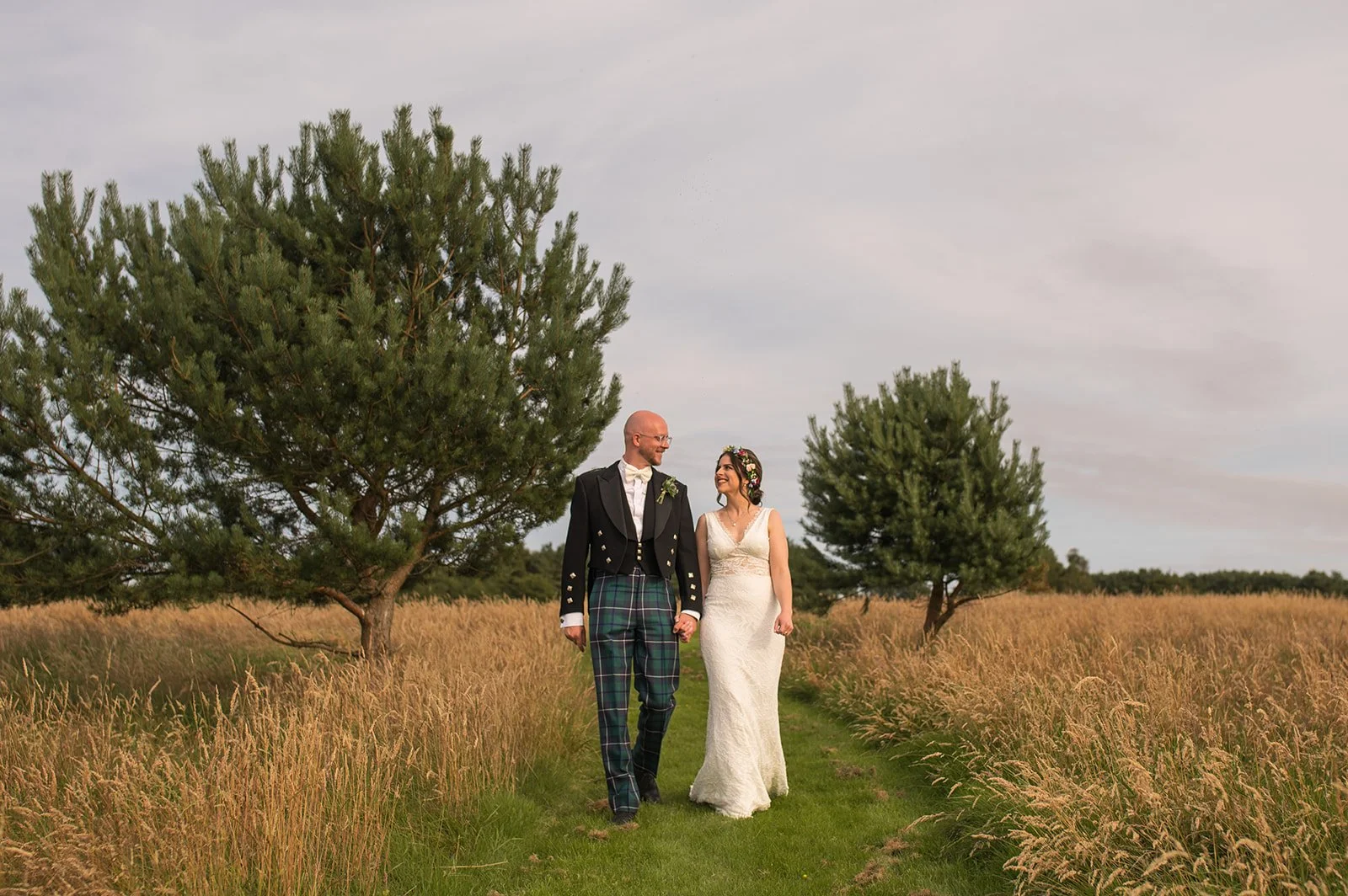A bride and groom walk hand in hand through a grassy field with tall grass, under a cloudy sky, surrounded by trees.