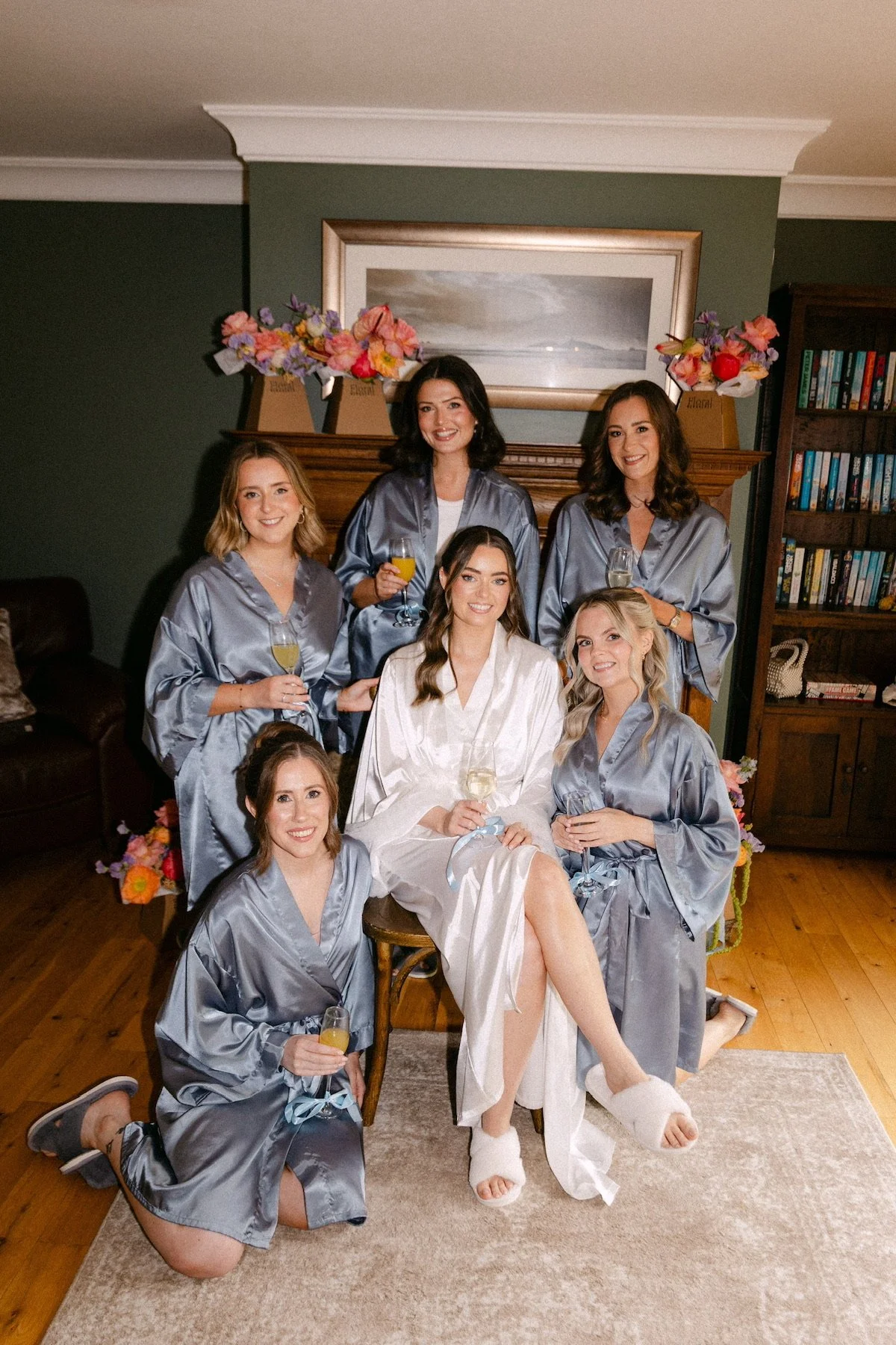 Six women gathered in a cozy living room, with one woman sitting in the center wearing a white robe, surrounded by five women in matching grey satin robes, all holding glasses of champagne or mimosa, celebrating a special occasion.