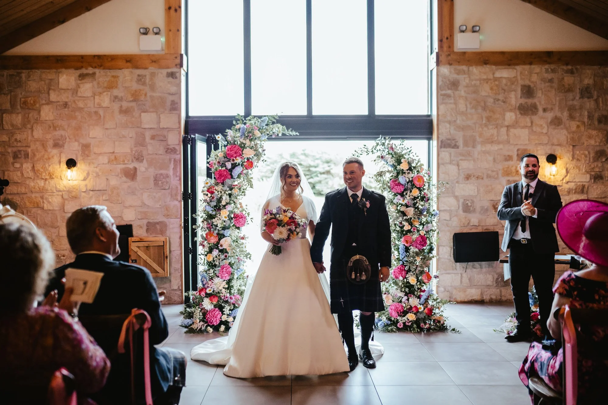 A bride and groom holding hands and smiling as they walk down the aisle during their wedding ceremony inside a rustic venue with stone and wood walls. The bride is wearing a white gown with a veil, holding a bouquet of flowers, and the groom is in a traditional Scottish kilt and jacket. Floral arrangements decorate the entrance.