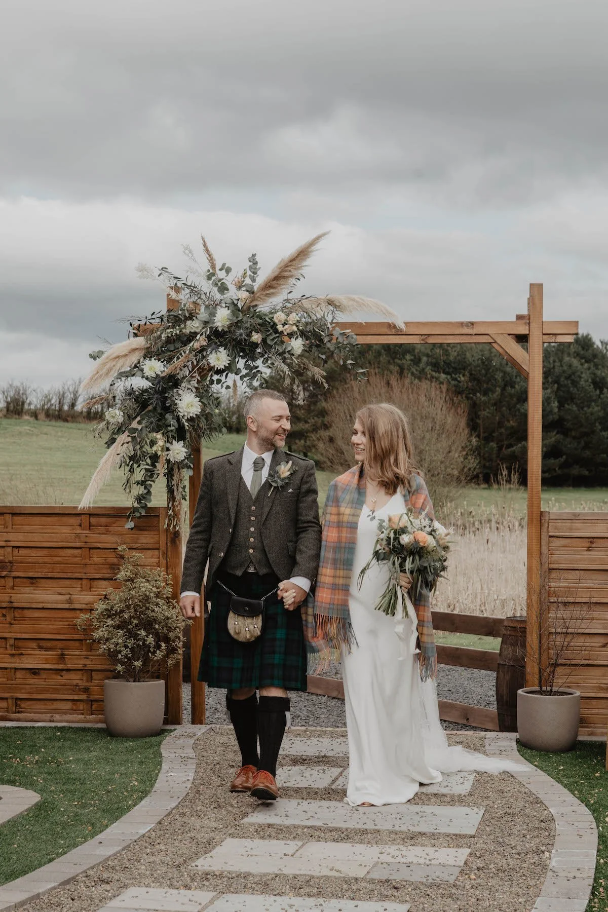 A couple, dressed in wedding attire, holding hands and smiling as they walk outside under a wooden arch decorated with flowers and pampas grass. The man is wearing a traditional Scottish kilt and jacket, and the woman is in a white wedding dress and a plaid shawl. They are on a stone and gravel pathway with potted plants around, with an open field and cloudy sky in the background.