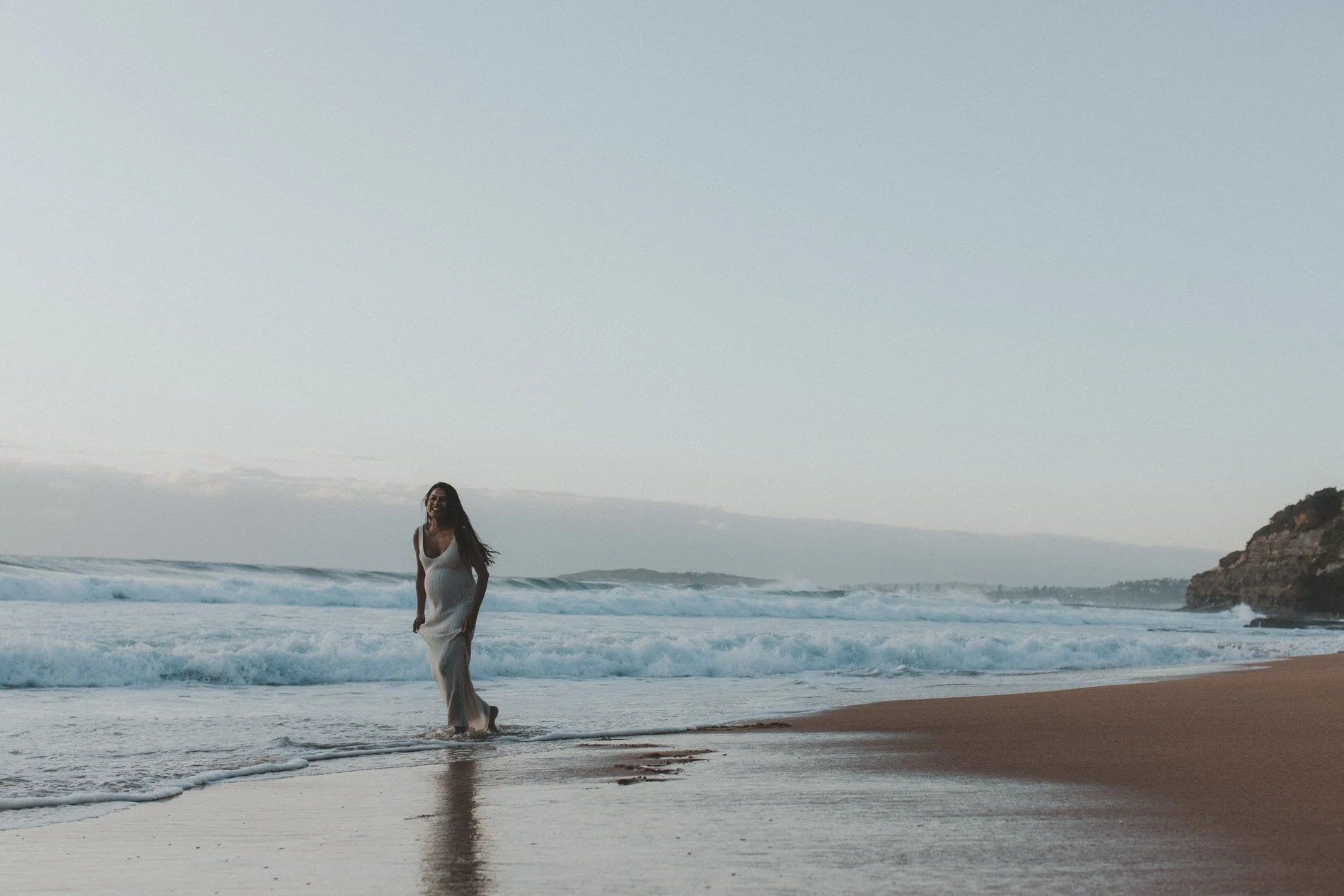 A woman in a white dress standing in the shallow surf at the beach during sunset, with cliffs and ocean waves in the background.