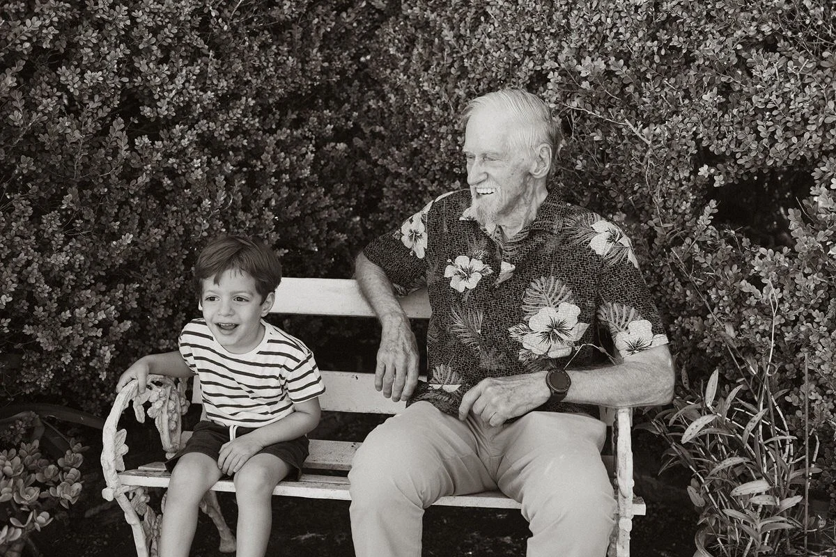 An elderly man and a young boy sitting on a bench outdoors surrounded by bushes. The boy is wearing a striped shirt and shorts, smiling and holding a rope. The man is wearing a floral shirt and khakis, smiling and looking at the boy.