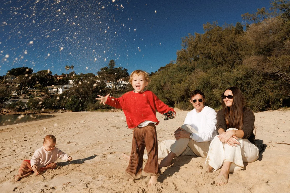 Two women and two children sitting on a sandy beach against a backdrop of trees and houses, with one child playing in the sand and the other holding a toy car, while the older child in red is throwing sand into the air.