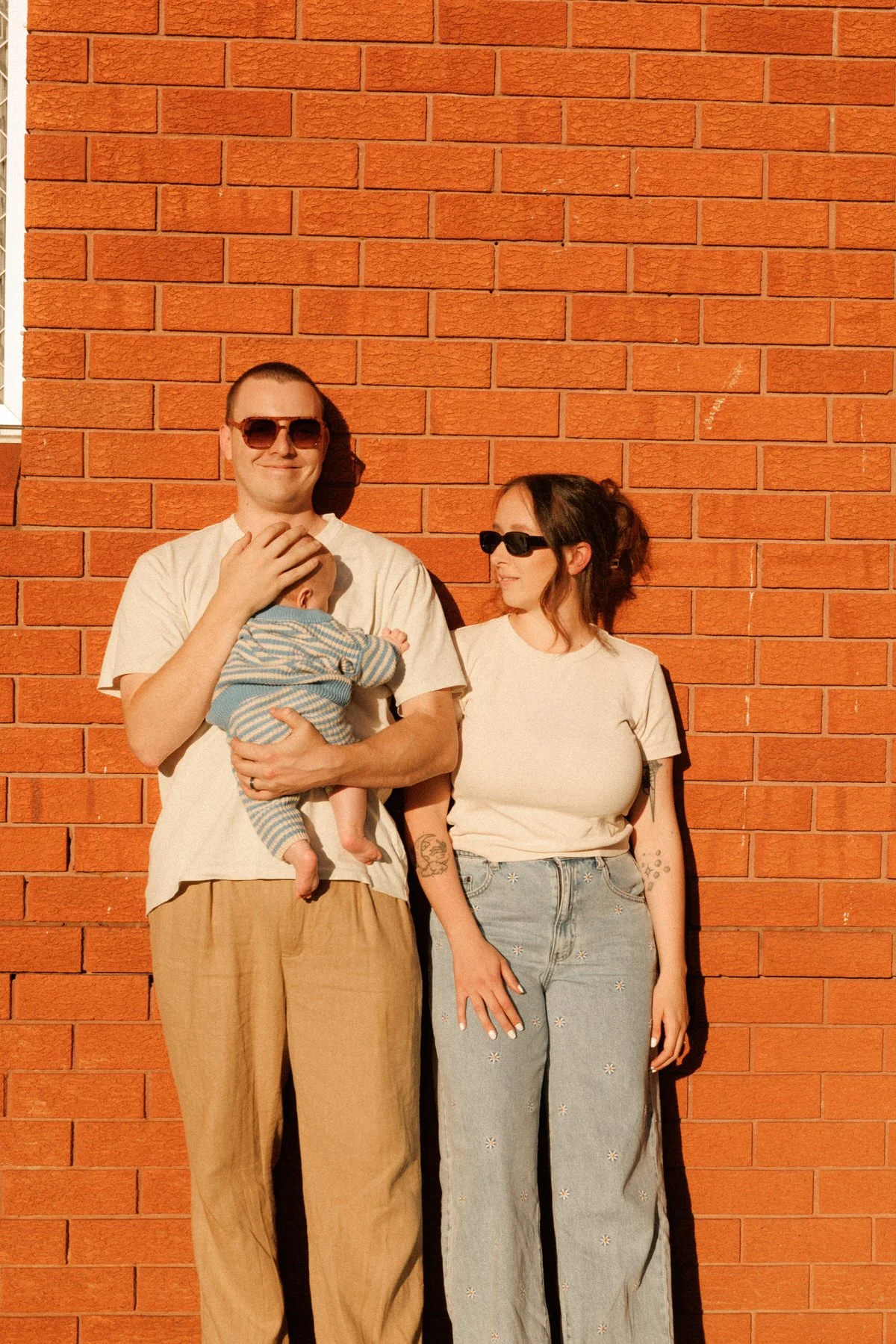 A man holding a baby and a woman standing next to him against a brick wall, all wearing sunglasses, with warm sunlight illuminating them.