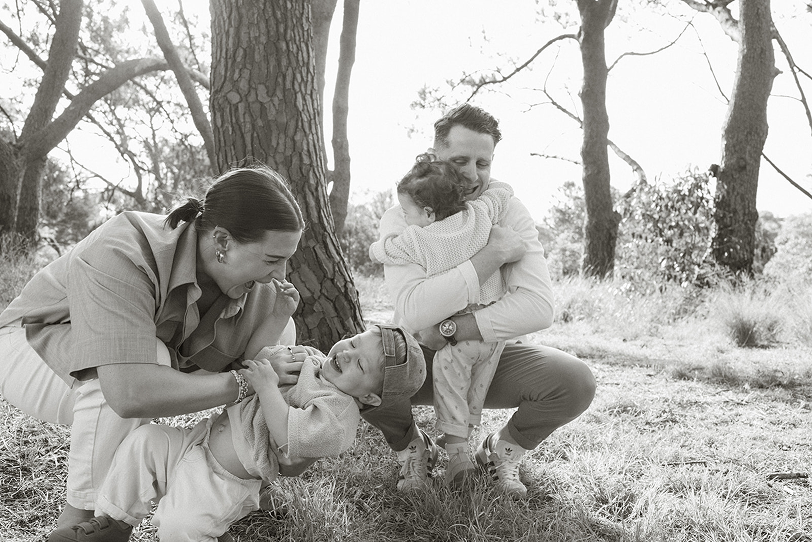 A family of four playing and laughing outdoors in a wooded area, with a woman and a young boy engaging in a playful tug-of-war and a man hugging a small child.