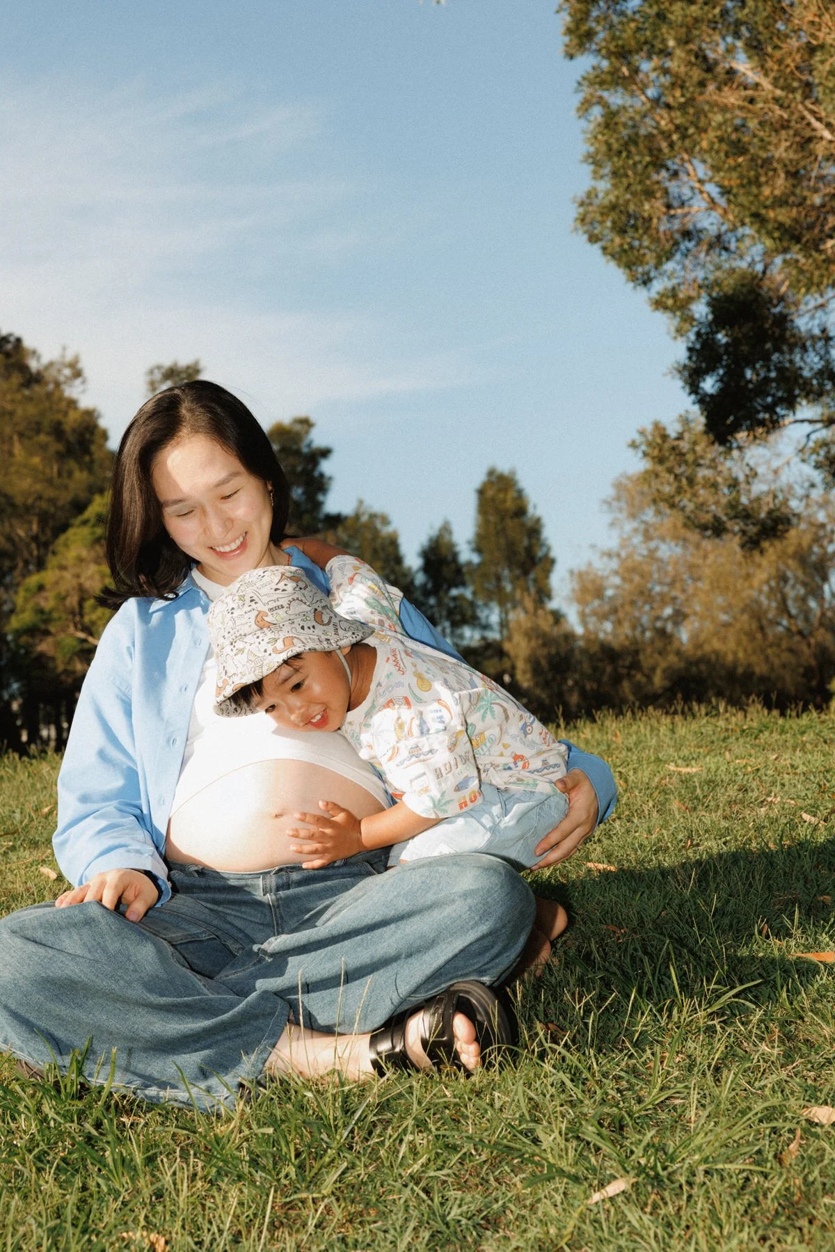A pregnant woman sitting cross-legged on the grass in a park, smiling with a young boy hugging her belly. The boy wears a hat and a patterned shirt, and they are enjoying sunny weather with trees and a blue sky in the background.