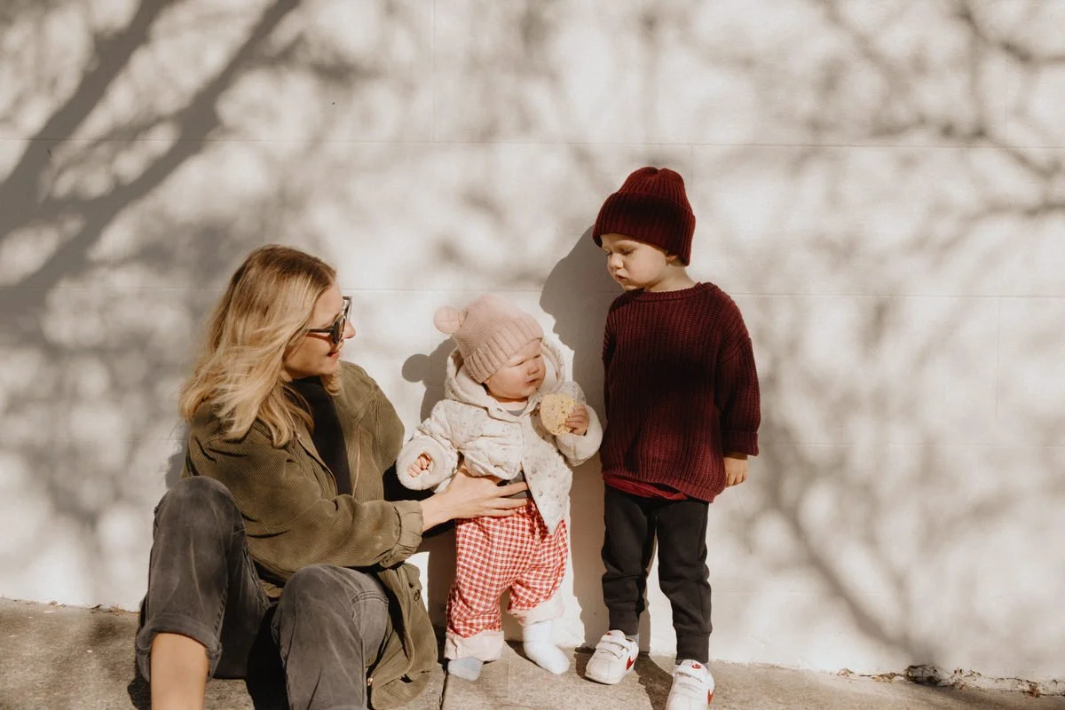 A woman sitting down next to two young children, one toddler and one young boy, against a white wall with shadows of tree branches, outdoors on a sunny day.
